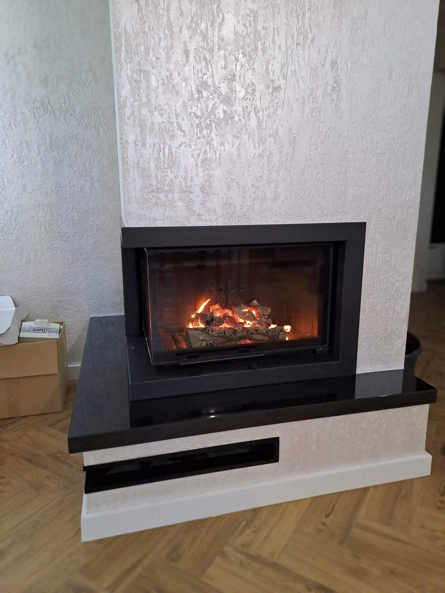 Nowoczesny kominek z czarną obudową i paleniskiem, ściana wykończona dekoracyjnym tynkiem strukturalnym, parkiet na podłodze.
