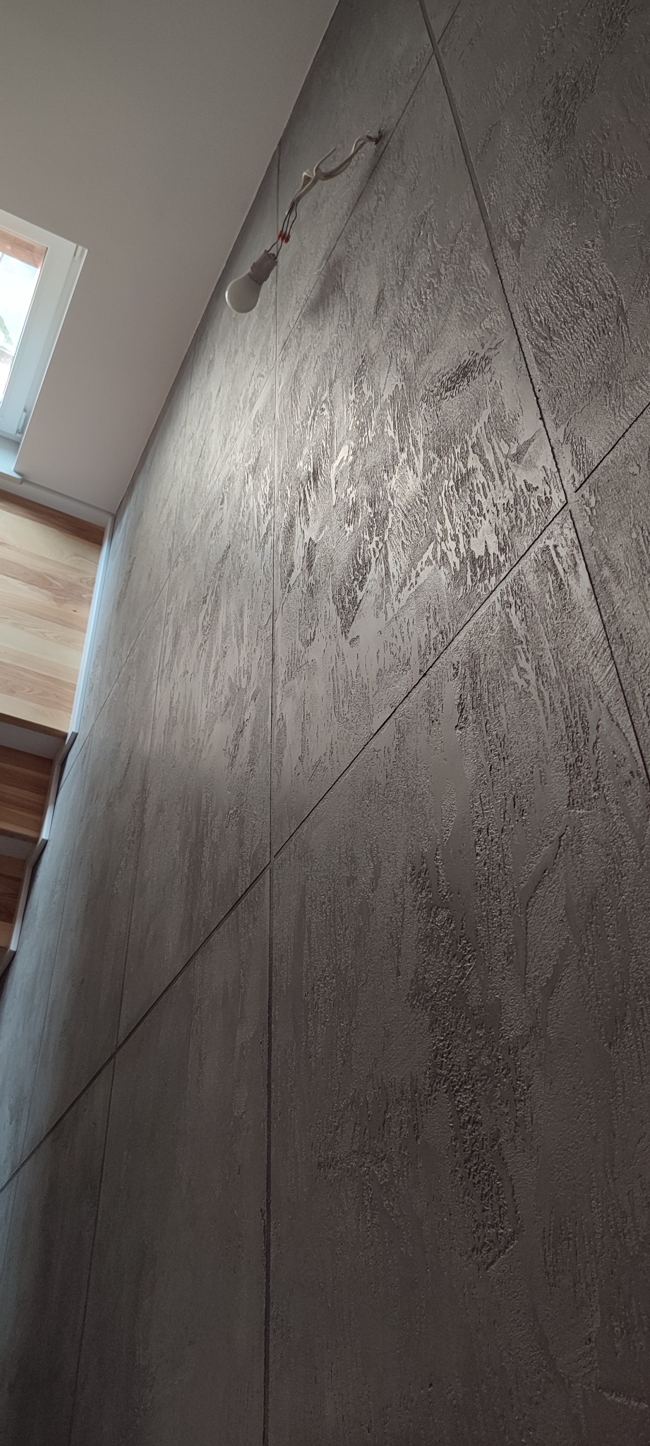 Ściana pokryta dekoracyjną, szarą fakturą imitującą beton, z widoczną żarówką na przewodach i fragmentem okna oraz drewnianych schodów w rogu.