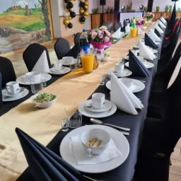 Uroczysty stół z czarnymi krzesłami, złoty bieżnik, biała zastawa, dekoracje kwiatowe, balony w tle, przygotowany na przyjęcie.