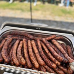 KONFIGURATOR IMPREZY Catering & Bankiet & Event www.konfiguratorimprezy.pl - Grillowane kiełbaski w metalowej brytfannie, gotowe do serwowania na imprezie plenerowej. W tle rozmyte postacie siedzące przy stołach.