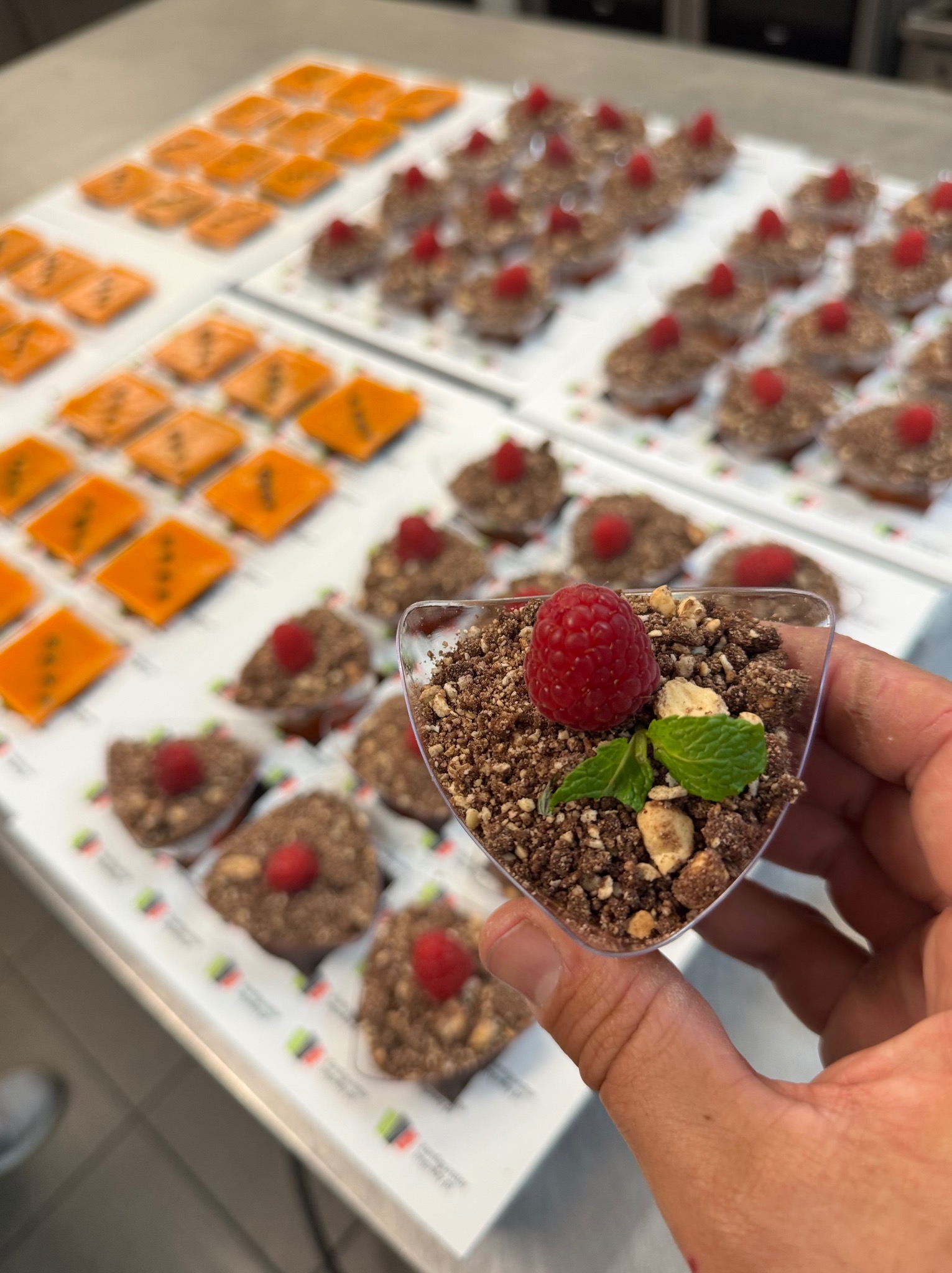 Deser w przezroczystym kubeczku, udekorowany maliną i miętą, trzymany w dłoni. W tle inne desery na białych tacach, w tym pomarańczowe i czekoladowe.