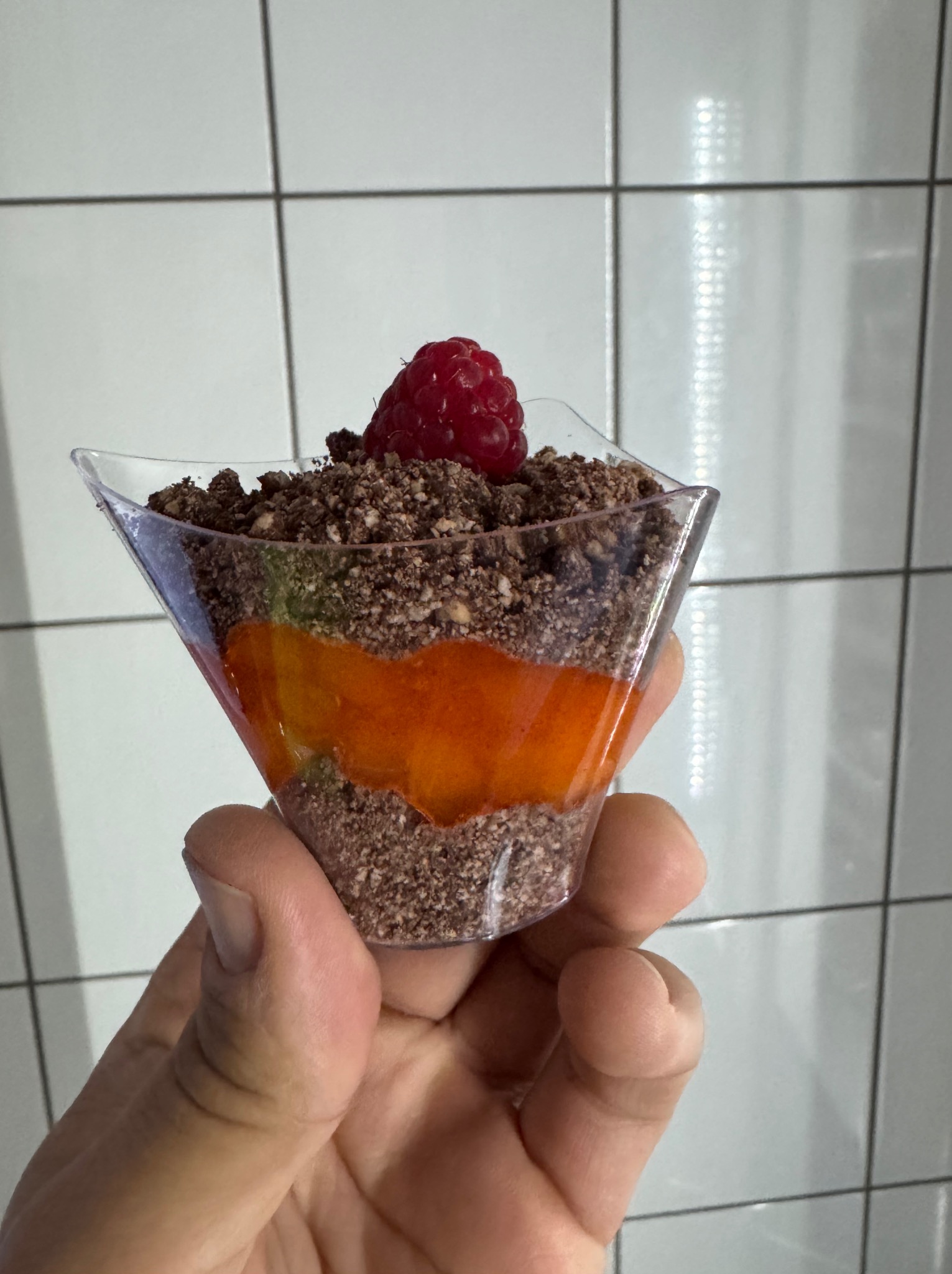 Deser w plastikowym kieliszku, warstwowy z kruszonką, musem pomarańczowym i maliną na wierzchu, trzymany w dłoni na tle białych płytek.