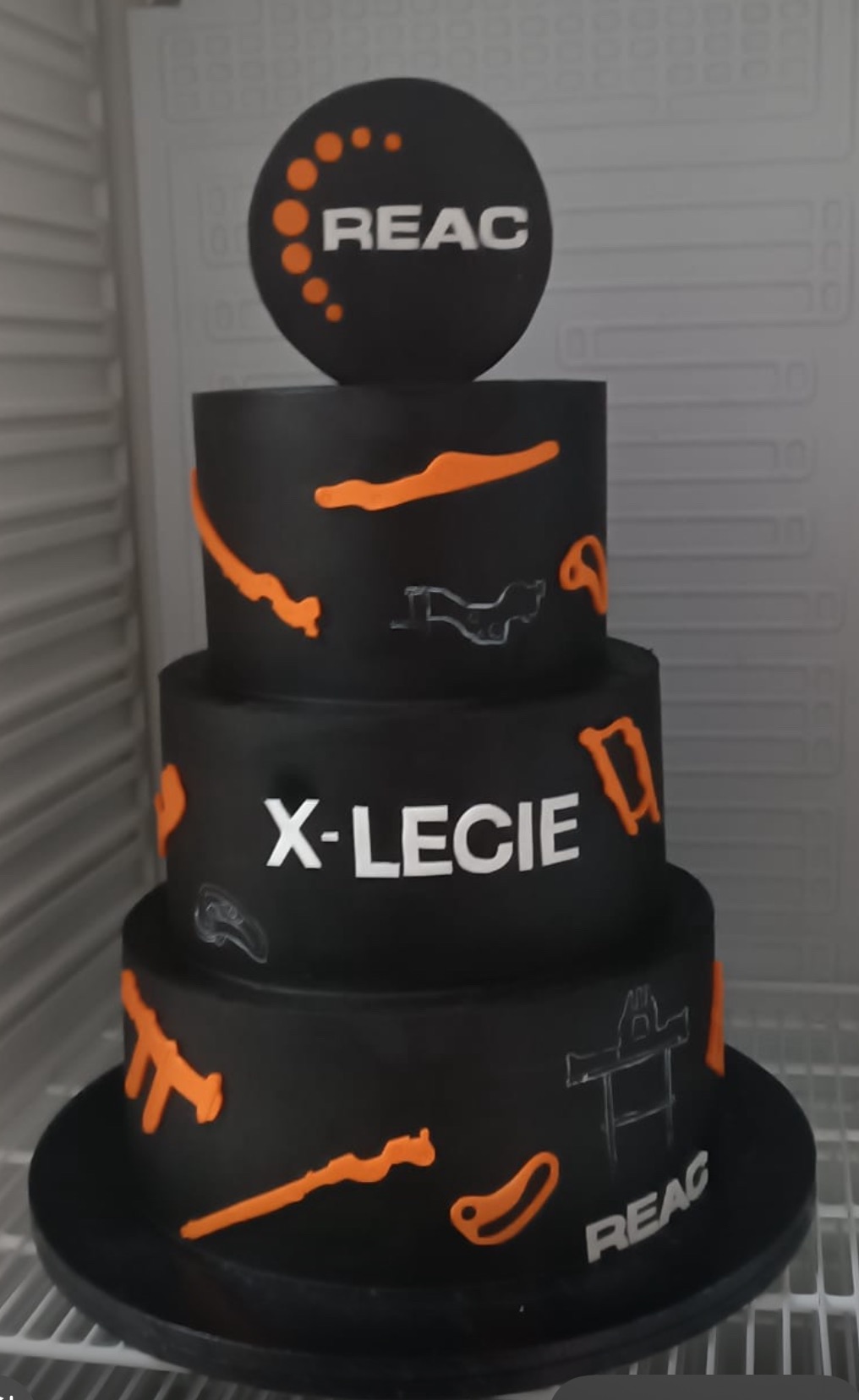 Czarny, trzypiętrowy tort z motywem korporacyjnym (logo REAC i napisem X-LECIE) oraz pomarańczowymi i szarymi elementami dekoracyjnymi. Nietypowy design.