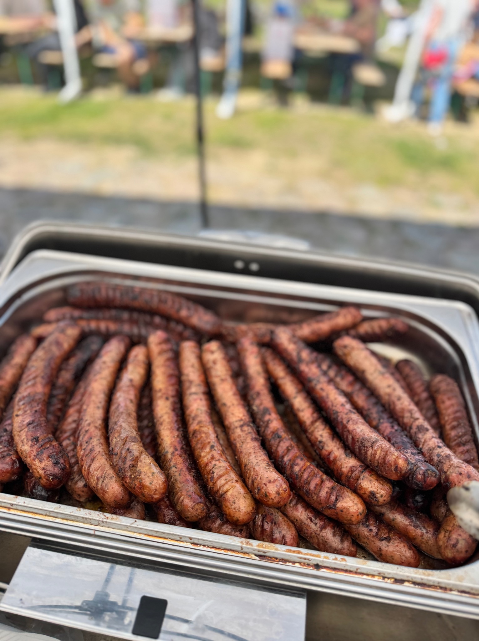 Grillowane kiełbaski w metalowej brytfannie, gotowe do serwowania na imprezie plenerowej. W tle rozmyte postacie siedzące przy stołach.