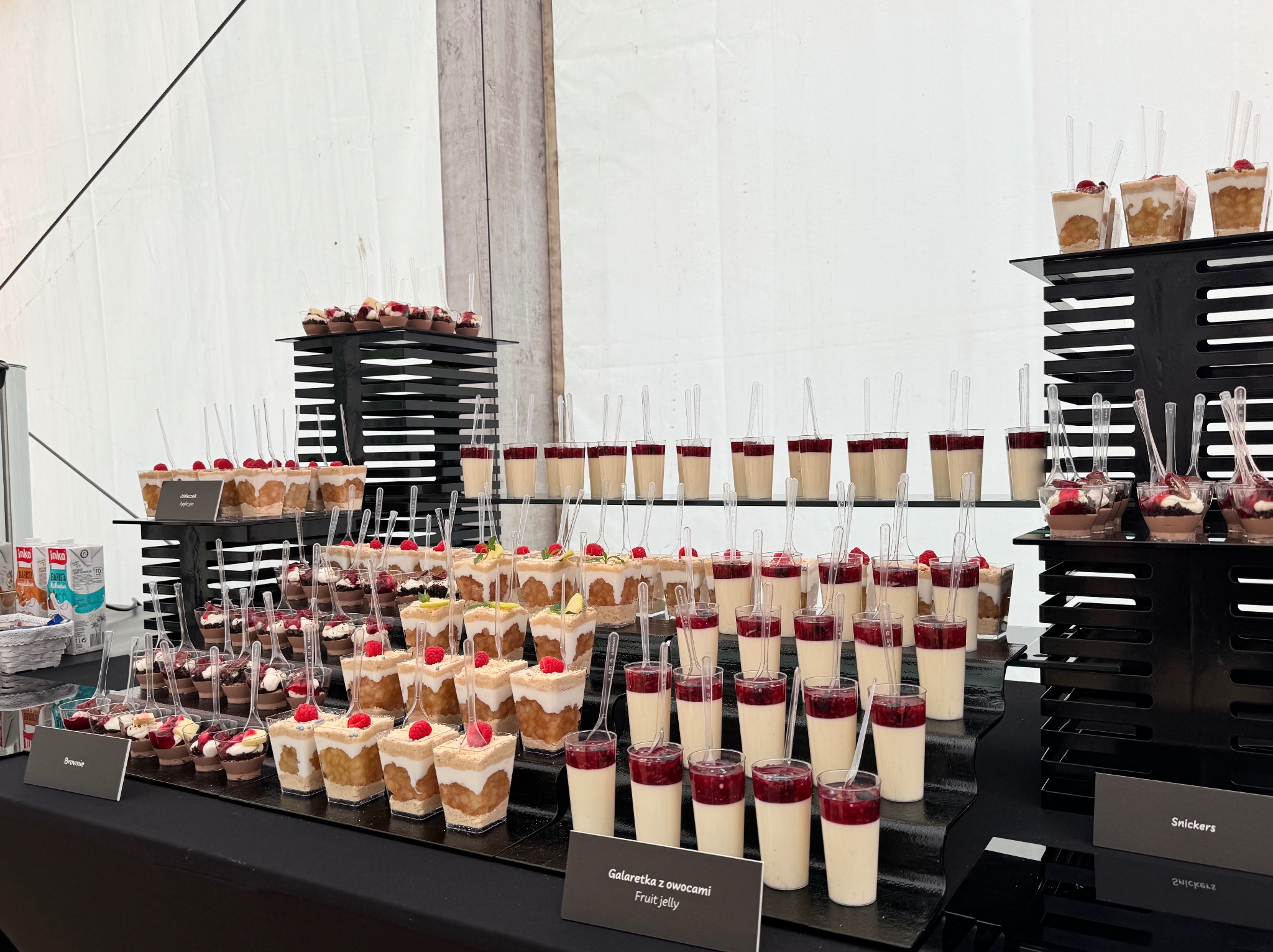 Stół cateringowy z deserami w szklankach: galaretka z owocami, brownie, jabłecznik, snickers. Desery na czarnych stojakach, z plastikowymi łyżeczkami.