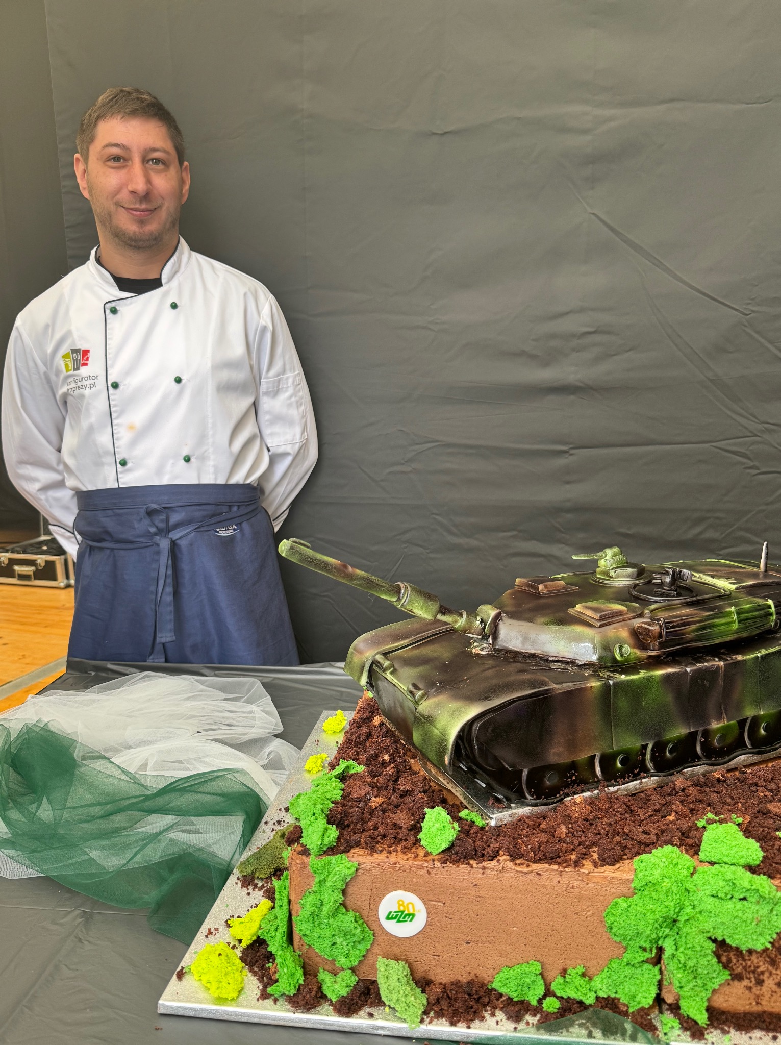 Uśmiechnięty cukiernik prezentuje tort w kształcie czołgu na tle ciemnej tkaniny. Czołg w barwach maskujących, na czekoladowym podłożu z zielonymi dekoracjami.