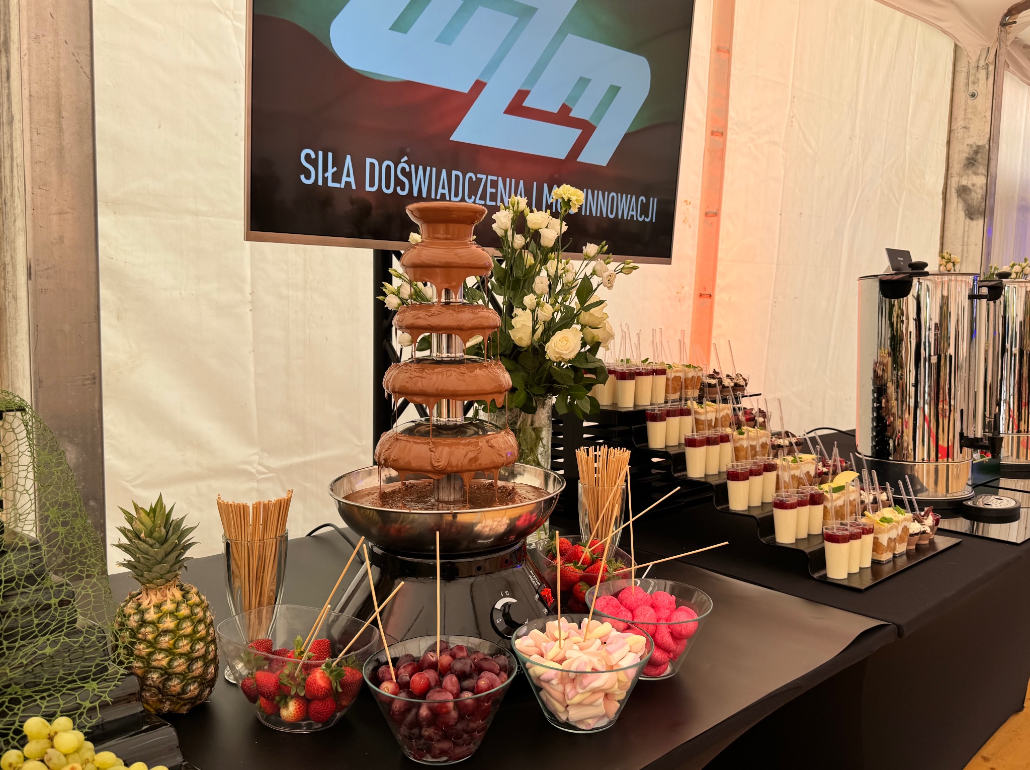 Fontanna czekoladowa otoczona owocami i deserami w szklankach, ustawiona na stole podczas eventu pod namiotem. Dekoracje kwiatowe i ananas.