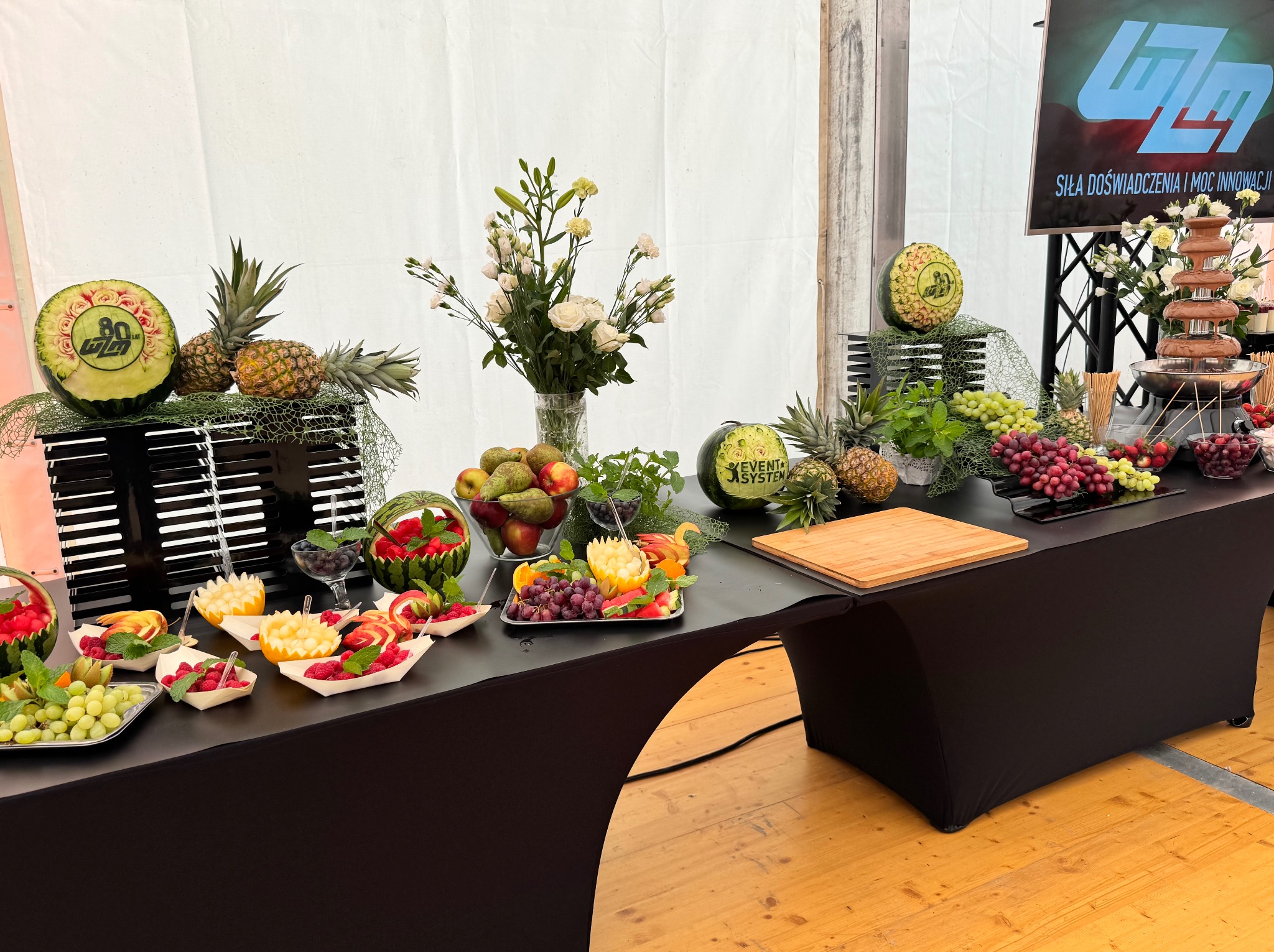 Stół bankietowy z dekoracjami owocowymi: rzeźbione arbuzy, ananasy, kwiaty i fontanna czekoladowa. Elegancki catering na przyjęcie.