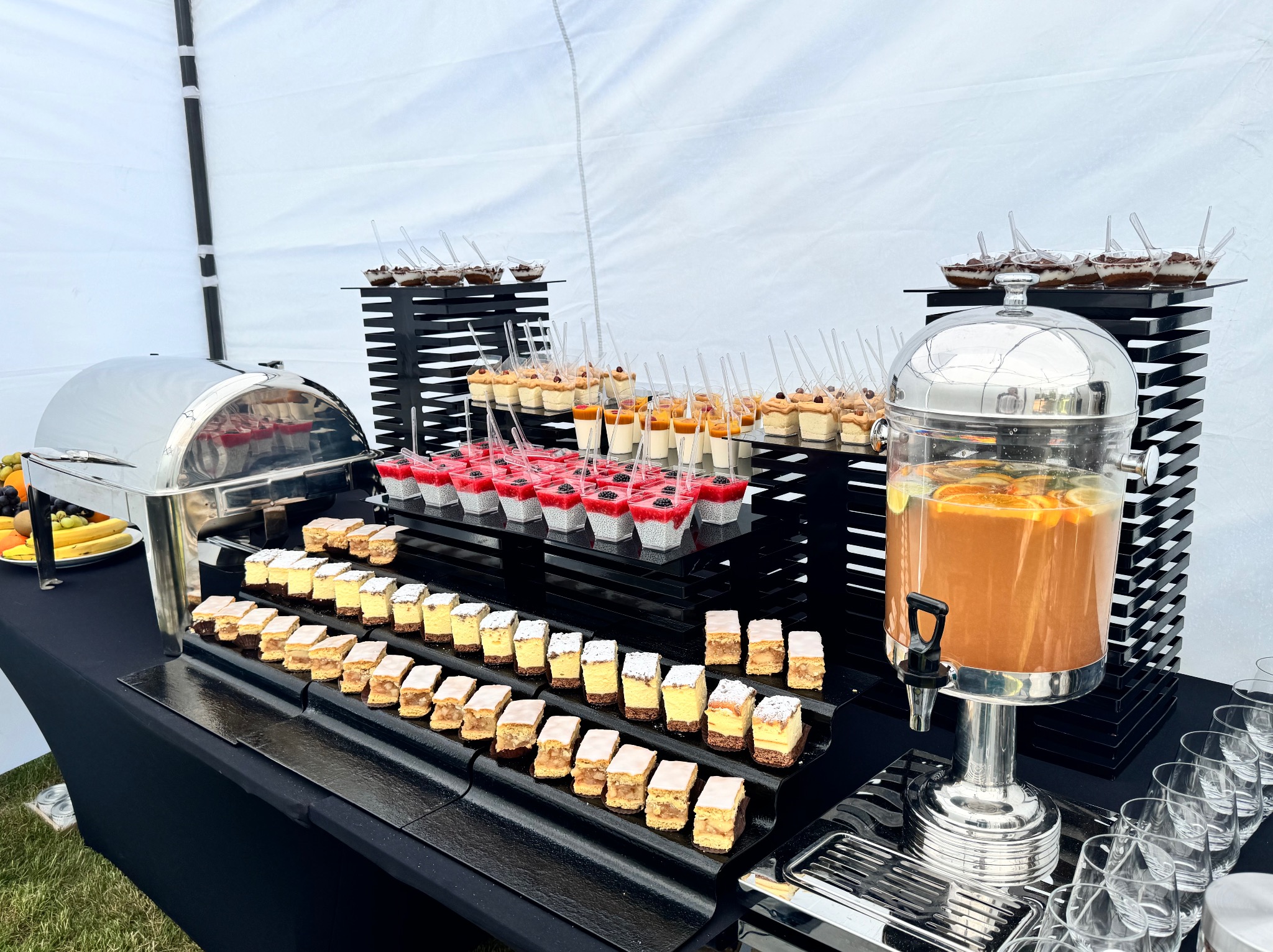 Stół cateringowy pod namiotem z deserami, napojem z pomarańczami i owocami. Eleganckie czarne podstawki, szklanki i naczynia ze stali nierdzewnej.