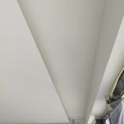 Nowoczesny sufit podwieszany z płyt kartonowo-gipsowych, widoczne prace wykończeniowe przy ścianach i zabezpieczone okno folią malarską.