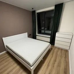 Minimalistyczna sypialnia z białym łóżkiem z materacem, komodą z szufladami i ciemnozielonymi zasłonami przy oknie, brązowa ściana w tle.