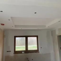 Wnętrze w trakcie prac wykończeniowych: sufit z podwieszaną konstrukcją i otworami na oświetlenie, ściany częściowo przygotowane do malowania, okno z widokiem na zieleń.