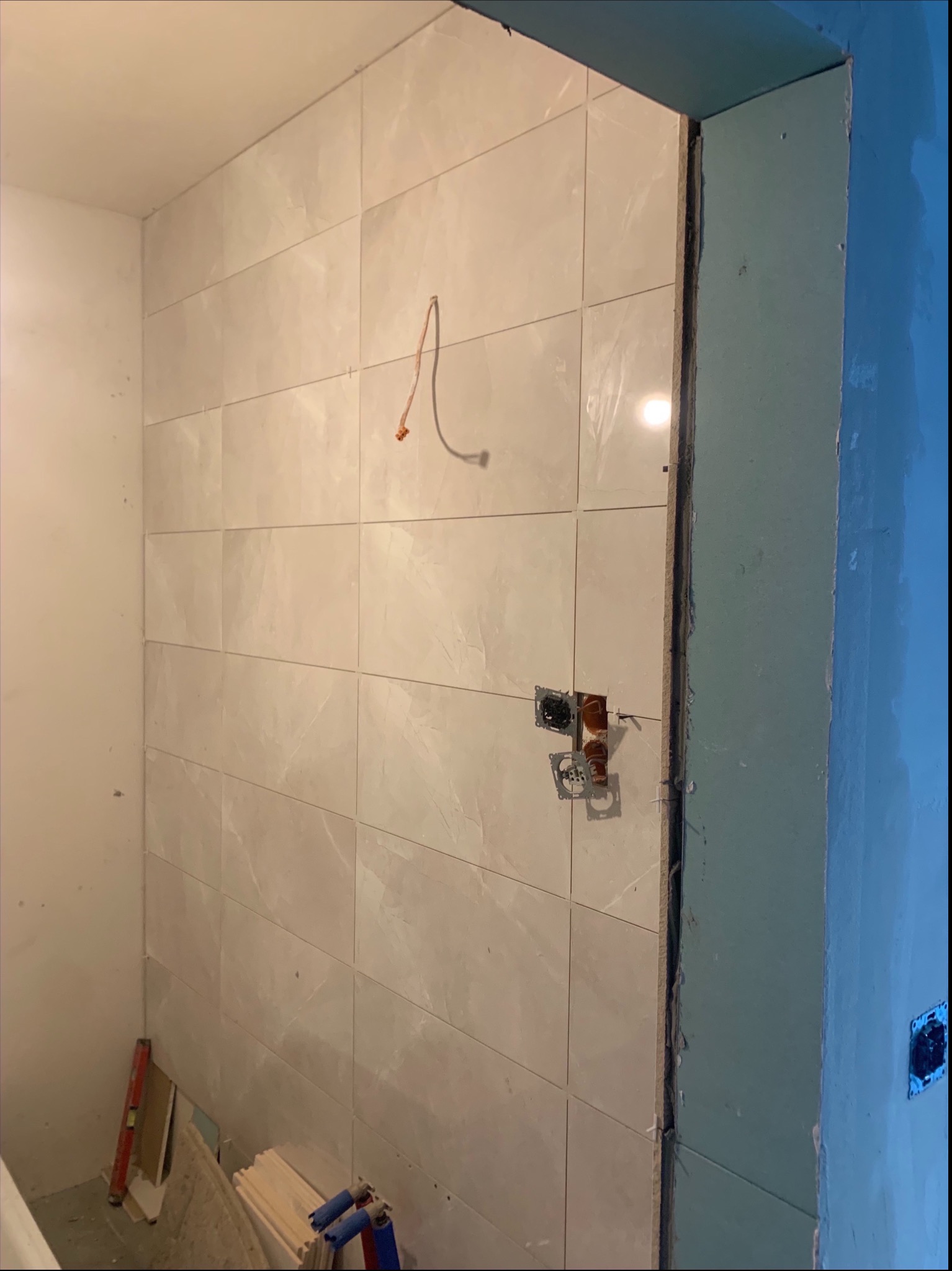 Ściana w łazience wyłożona jasnymi, kwadratowymi płytkami ceramicznymi, z widocznymi puszkami elektrycznymi bez osprzętu, przewodem zwisającym ze ściany i fragmentem ściany z płyty...