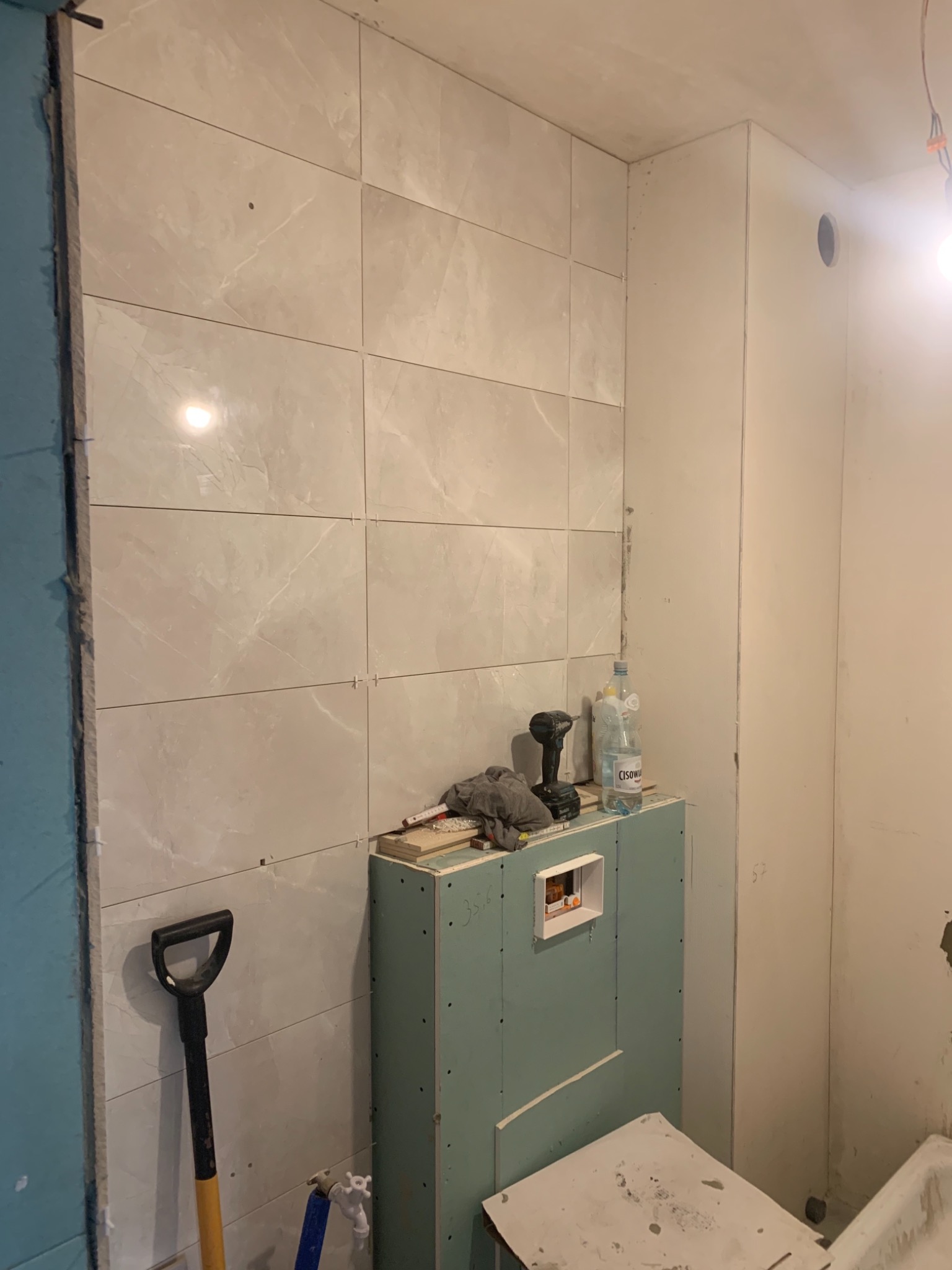 Ściana łazienki w trakcie wykańczania: ułożone jasne płytki imitujące marmur, obudowa stelaża WC z płyty karton-gips, widoczne narzędzia, butelka wody i poziomica.