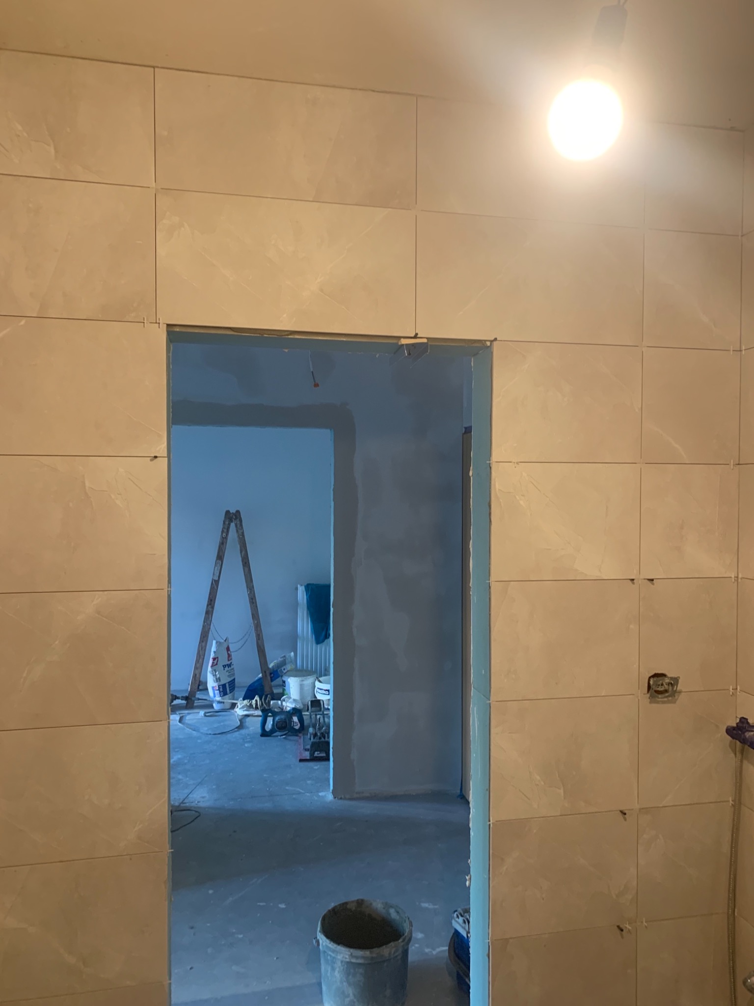 Ściana łazienki w trakcie układania beżowych płytek z marmurkowym wzorem, widoczna drabina i narzędzia w przejściu do innego pomieszczenia, surowe wnętrze.