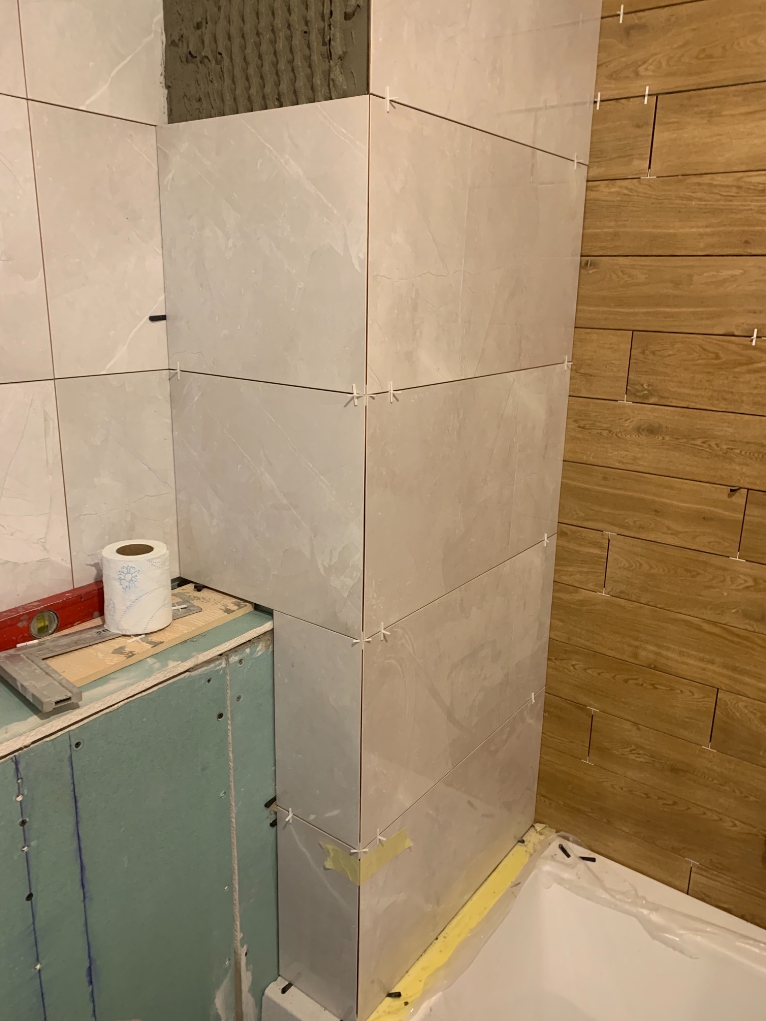 Świeżo ułożone, jasne płytki imitujące marmur na ścianie łazienki, z widocznymi krzyżykami dystansowymi i fragmentem ściany z okładziną drewnopodobną; poziomica i rolka papieru toaletowego...