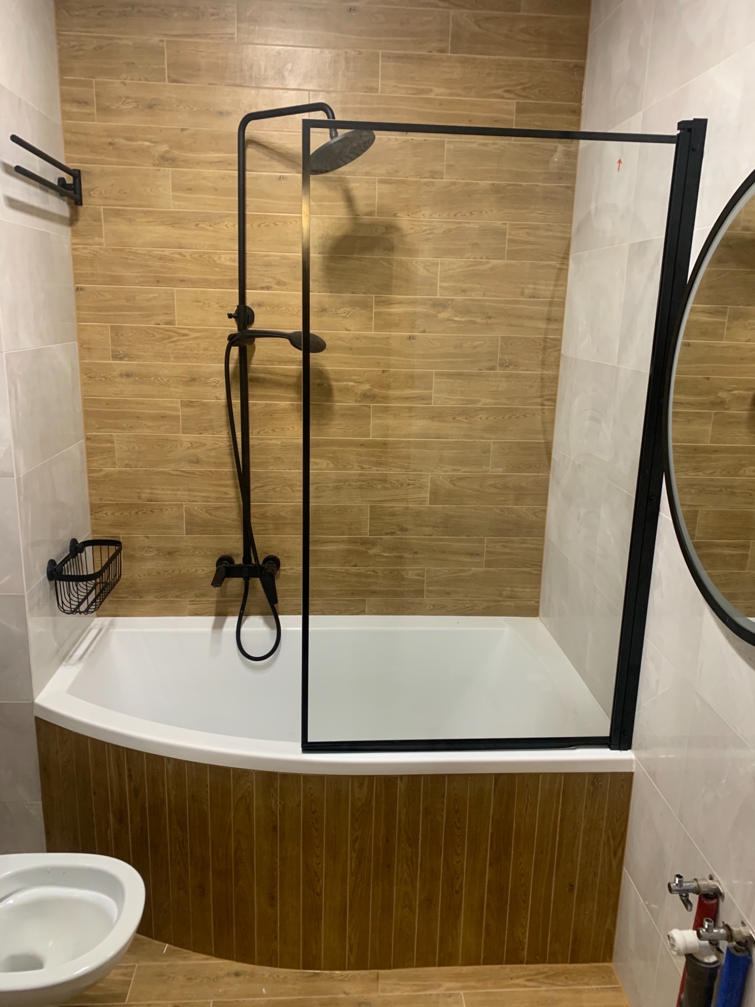 Wykończona łazienka z wanną, kabiną prysznicową z czarną ramą, armaturą w kolorze czarnym, drewnopodobnymi płytkami na ścianach i obudowie wanny oraz częściowo widoczną toaletą.