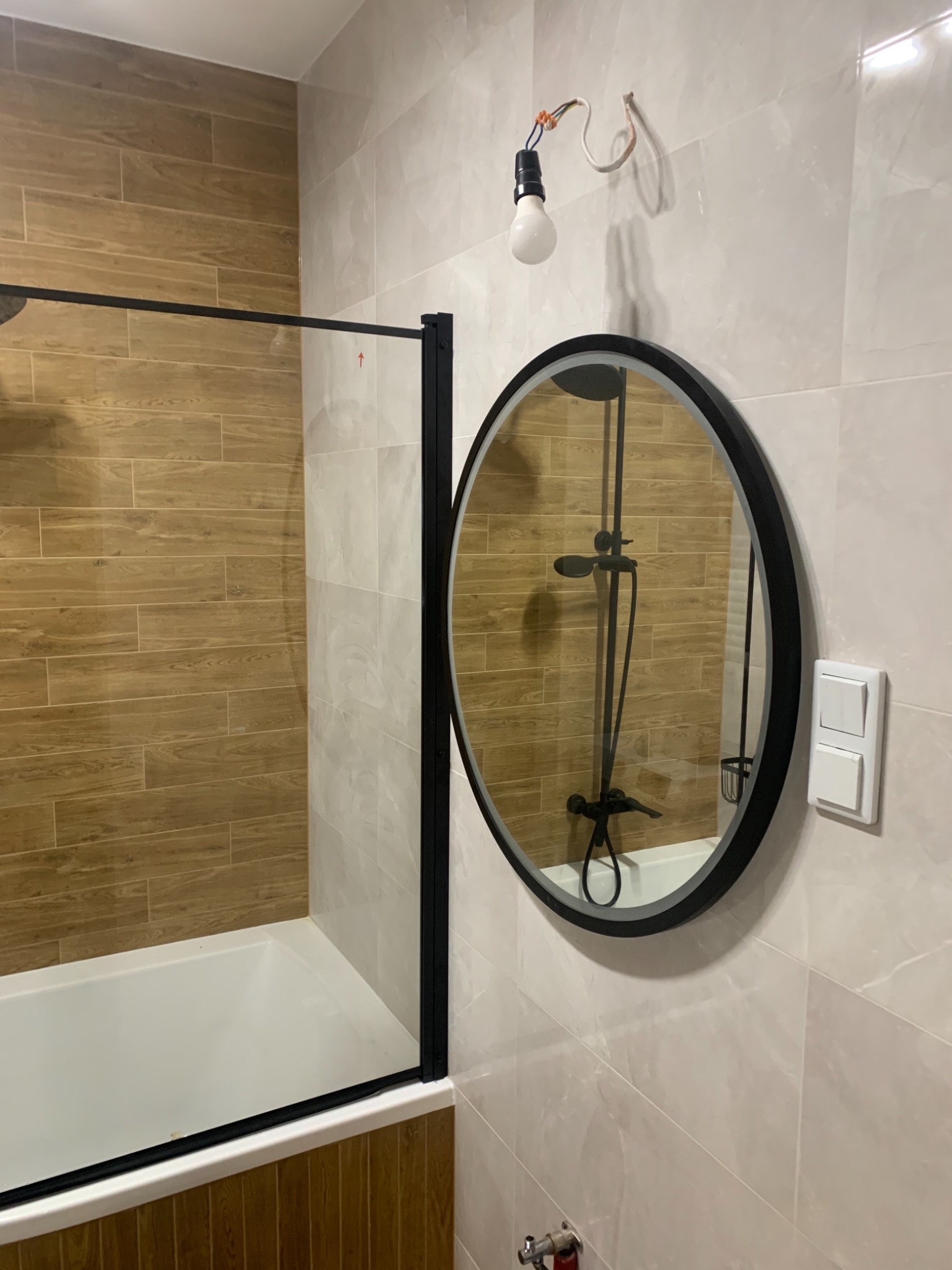 Wykończona łazienka z wanną z czarnym parawanem, okrągłym lustrem z czarną ramą odbijającym baterię wannową i prysznicową, oraz niedokończonym oświetleniem sufitowym z widocznymi kablami.