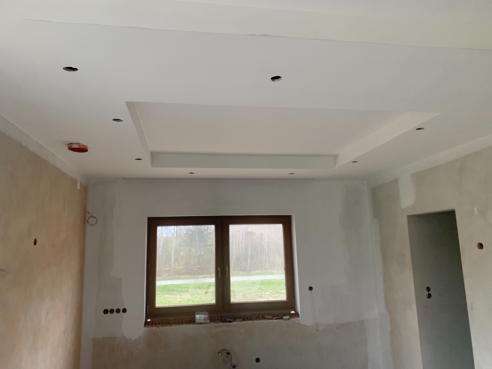 Wnętrze w trakcie prac wykończeniowych: sufit z podwieszaną konstrukcją i otworami na oświetlenie, ściany częściowo przygotowane do malowania, okno z widokiem na zieleń.