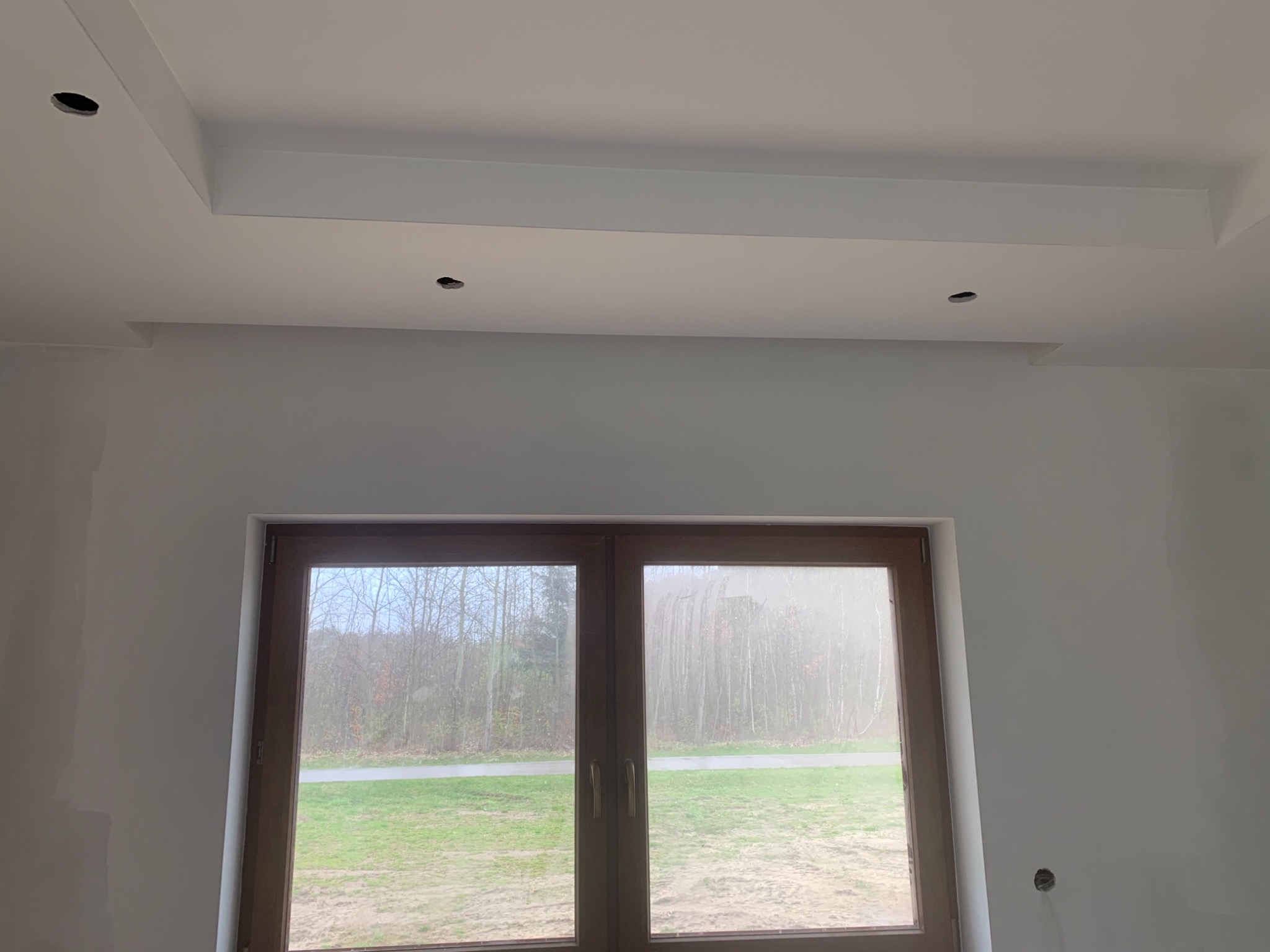 Wnętrze pokoju po remoncie, widoczne świeżo pomalowane ściany w kolorze białym, drewniane okno z widokiem na zewnątrz oraz sufit z zamontowanymi elementami dekoracyjnymi i otworami na oświetlenie.