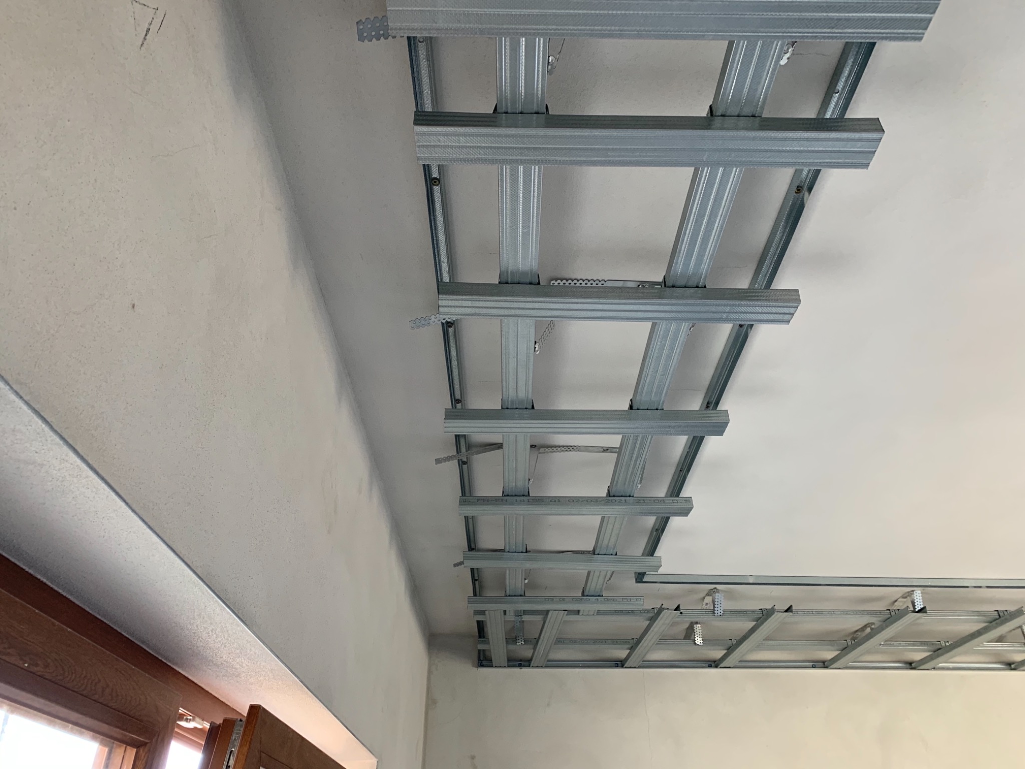 Metalowa konstrukcja sufitu podwieszanego, widoczna od dołu, z elementami mocującymi i profilem przy oknie, przygotowana do montażu płyt.