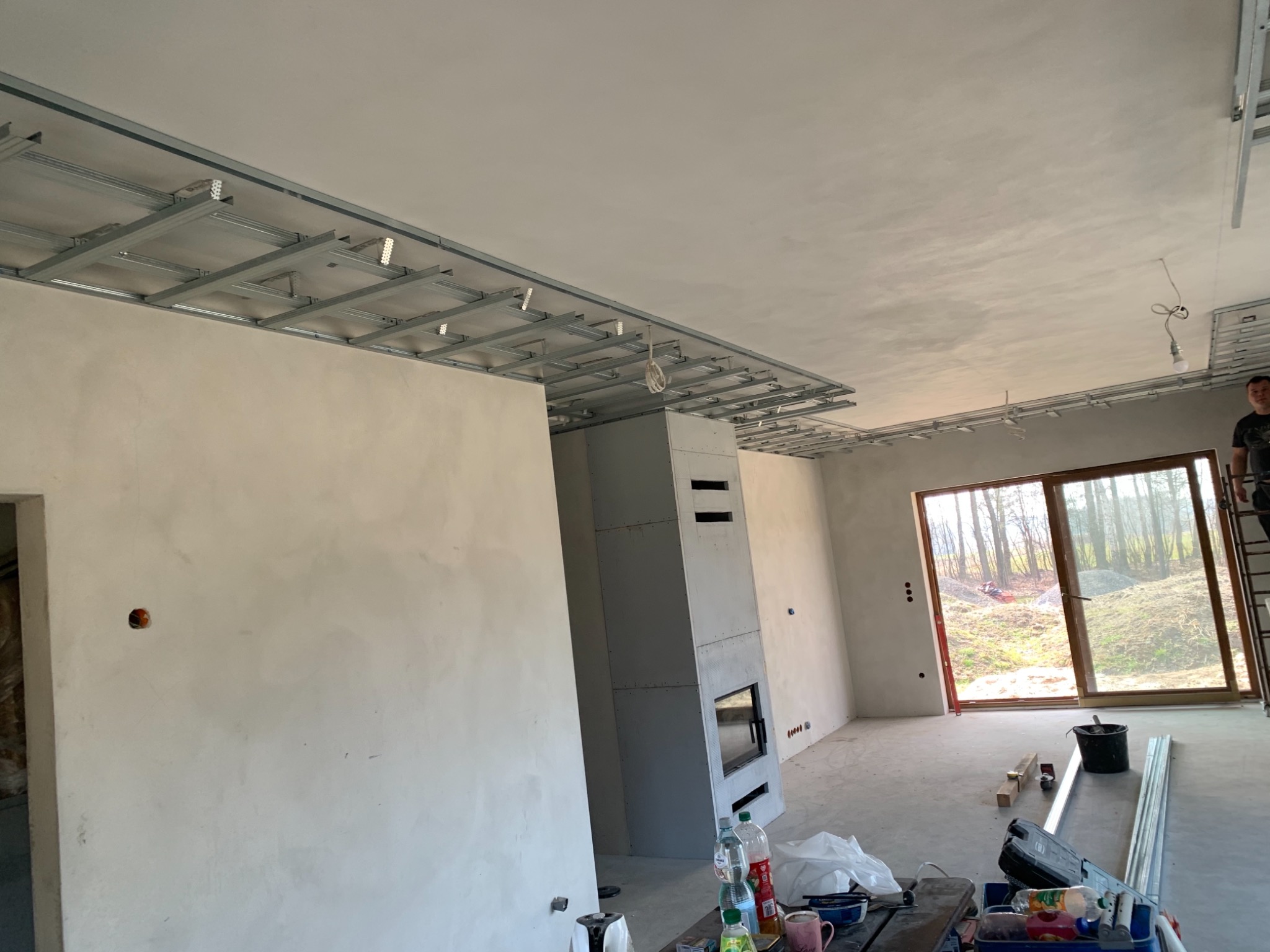 Wnętrze w trakcie wykańczania: widoczne metalowe profile sufitowe, ściany po nałożeniu gładzi, obudowa kominka z płyty karton gips, narzędzia i materiały budowlane na podłodze, oraz częściowo...