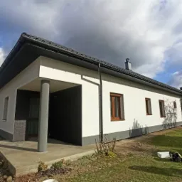 Jednopiętrowy dom z białymi ścianami, ciemną dachówką i szarymi elementami wykończeniowymi, widoczny z boku, z elementami zielonego trawnika w tle.