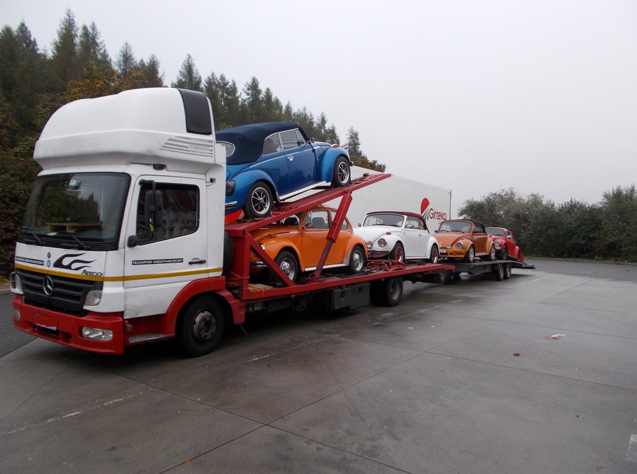 Laweta transportuje kolekcję zabytkowych Volkswagenów Garbusów w różnych kolorach. Widoczny biały ciągnik siodłowy z czerwoną ramą i logo Girteka na naczepie.