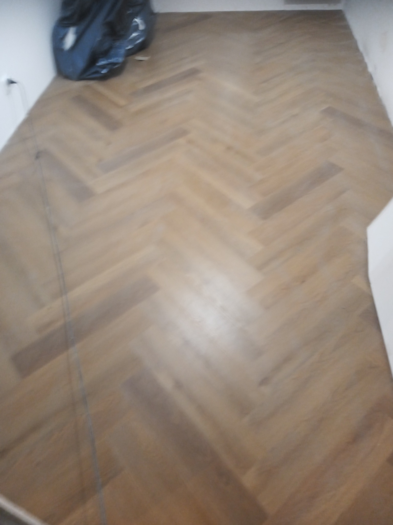 Ułożony parkiet w jodełkę, widoczny worek z odpadami w rogu pokoju, jasne ściany.