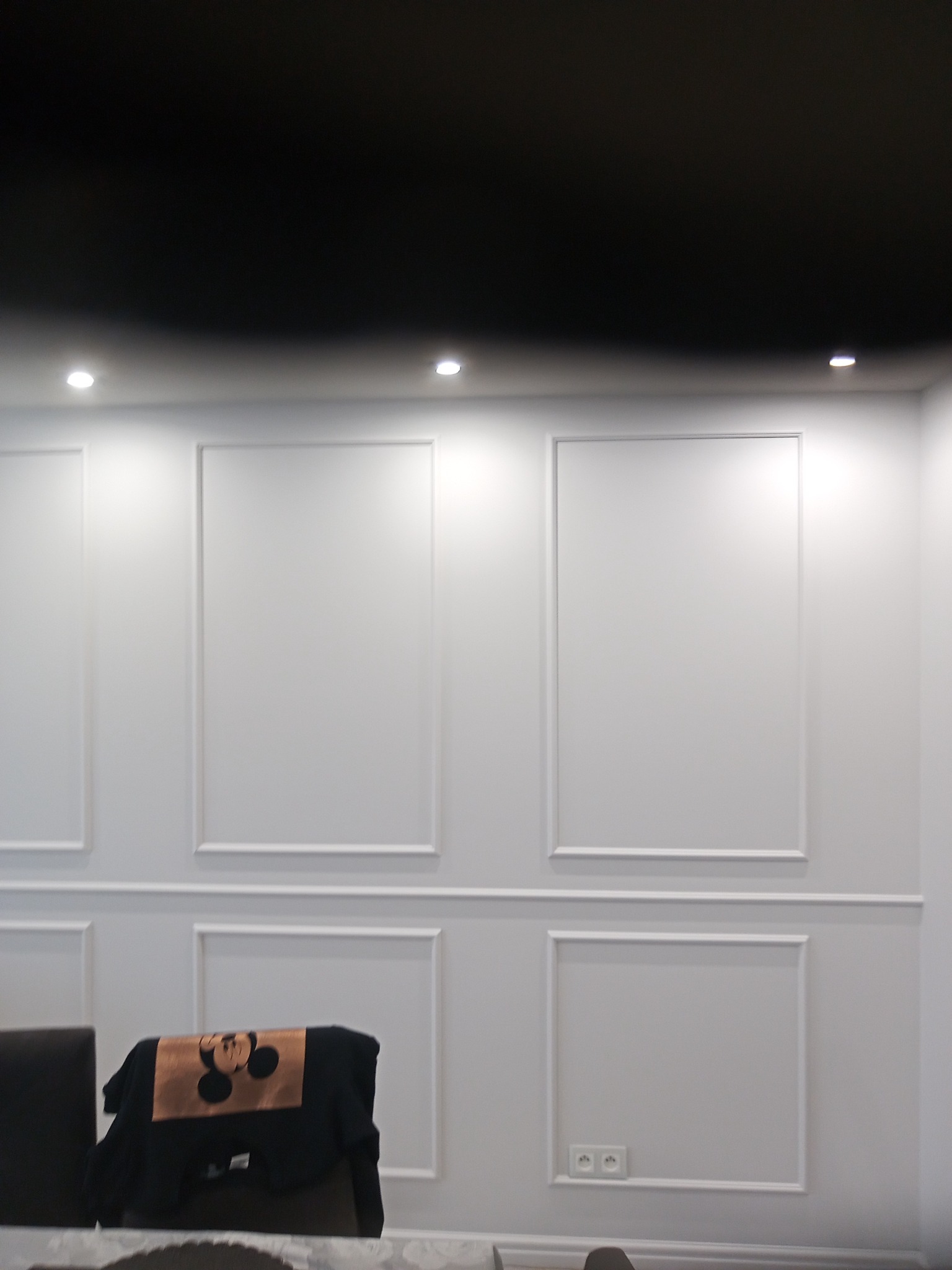 Biała ściana z dekoracyjnymi, symetrycznymi panelami w stylu klasycznym, czarny sufit z punktowym oświetleniem, widoczne gniazdka elektryczne i fragment krzesła z bluzą z motywem Myszki Miki.