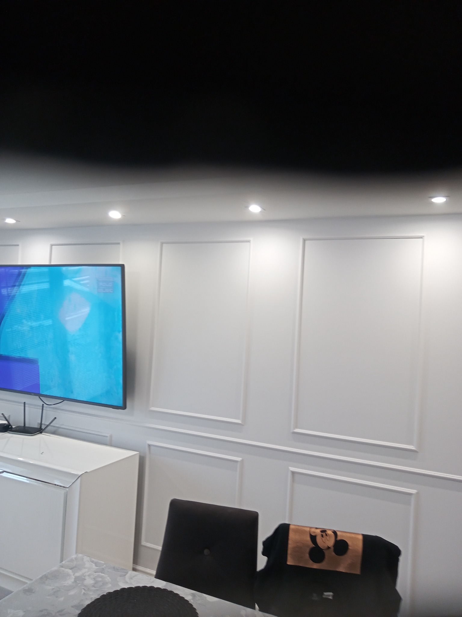 Nowoczesne wnętrze z białymi ścianami zdobionymi sztukaterią, duży telewizor LCD na ścianie, biała komoda i stół z szarym blatem, krzesła w odcieniach szarości, oświetlenie punktowe sufitowe.