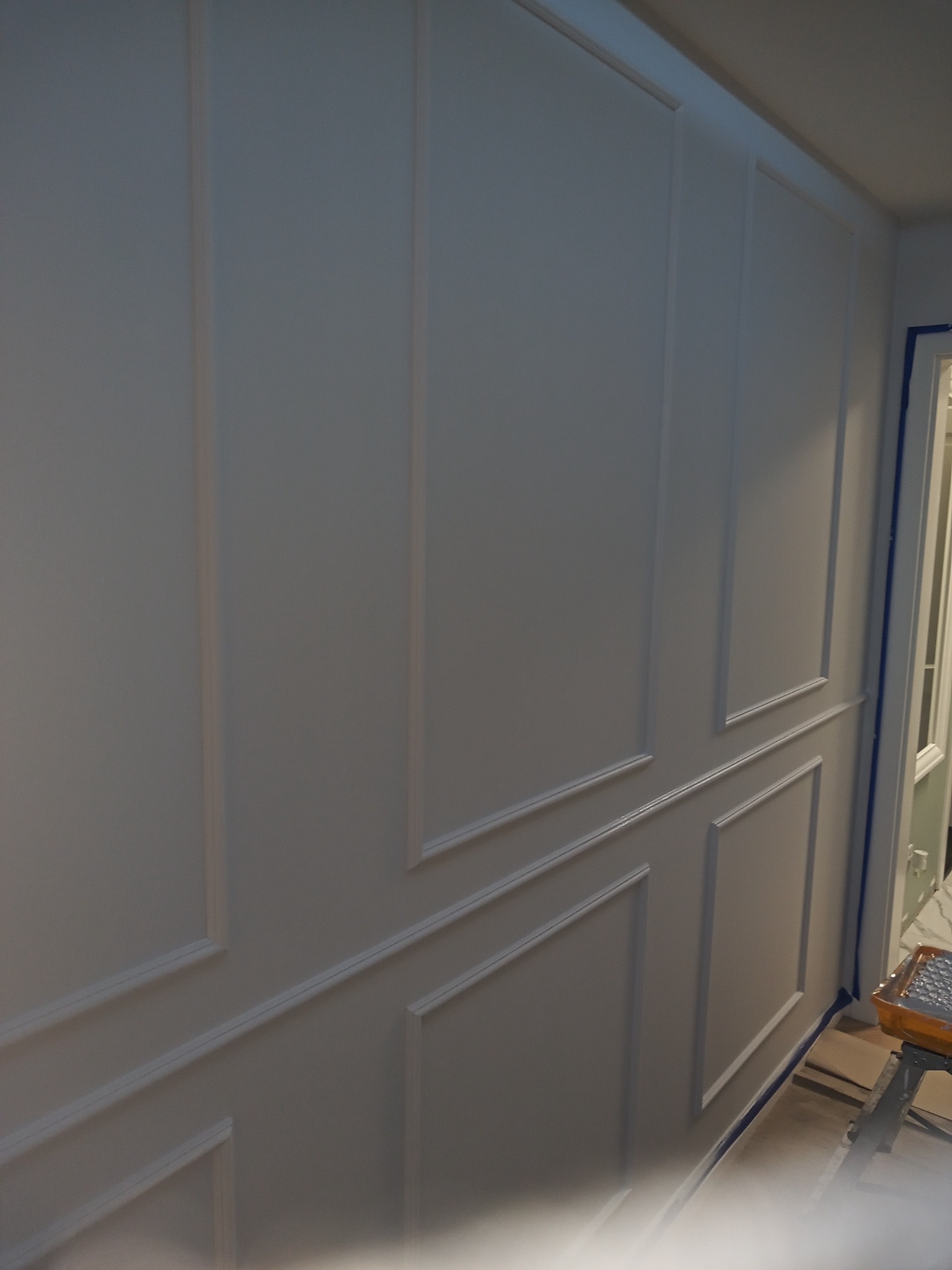 Ściana z dekoracyjnymi listwami pomalowana na szaro, zabezpieczona taśmą malarską przy drzwiach i podłodze, widoczny fragment drabiny malarskiej.