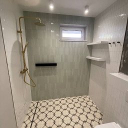 GROSS-CHEF PATRYCJA ŁABSZ - Wykończona łazienka w Nowej Woli z kabiną prysznicową z złotą armaturą, geometryczną podłogą w romby i pionowymi płytkami w odcieniach szarości na ścianach, uzupełniona o białe półki i okno.