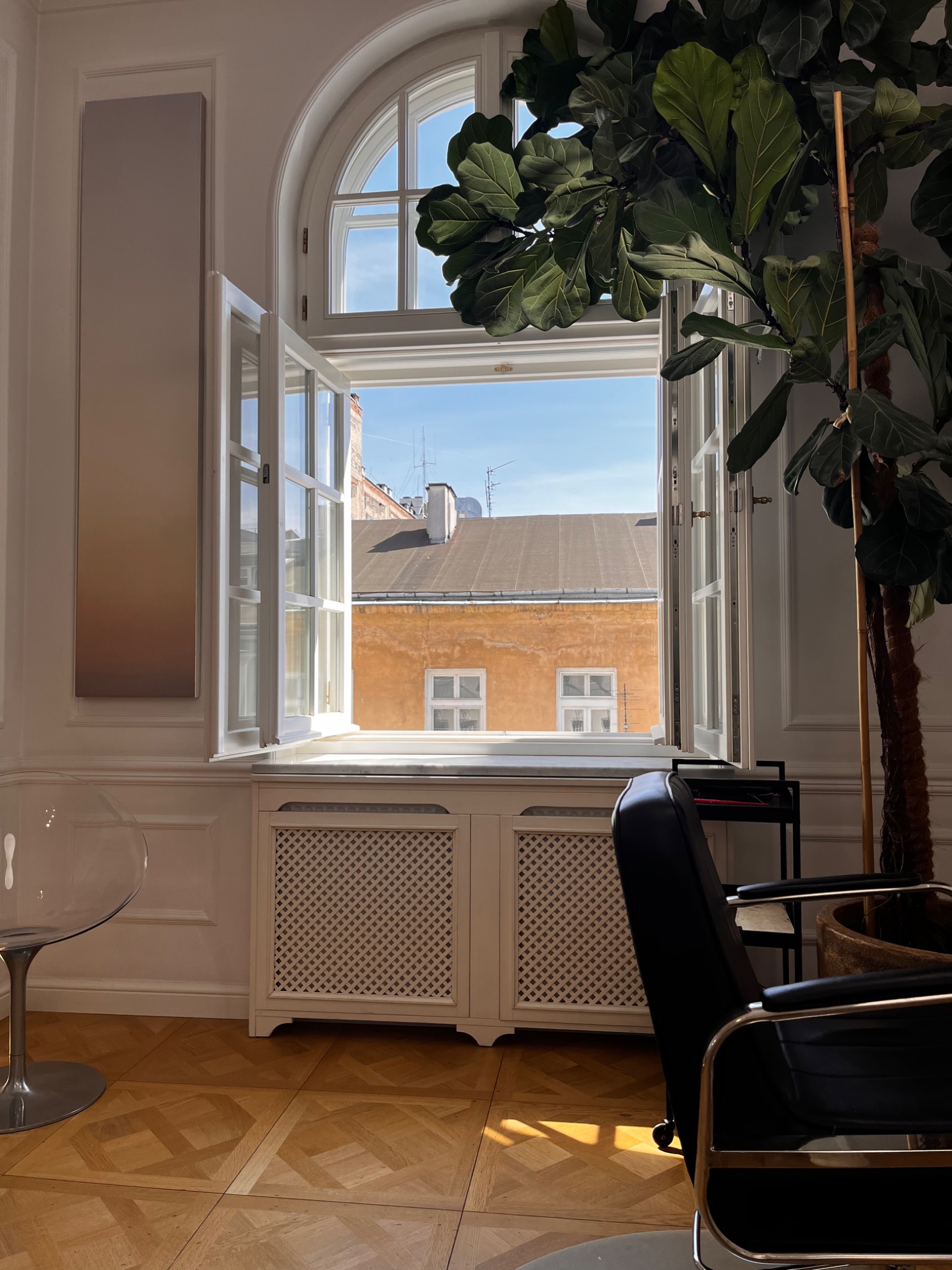 Jasne wnętrze z otwartym oknem łukowym, przez które widać budynek; parkiet w geometryczny wzór, białe ściany z dekoracyjnymi listwami, duża roślina doniczkowa i designerskie krzesło.