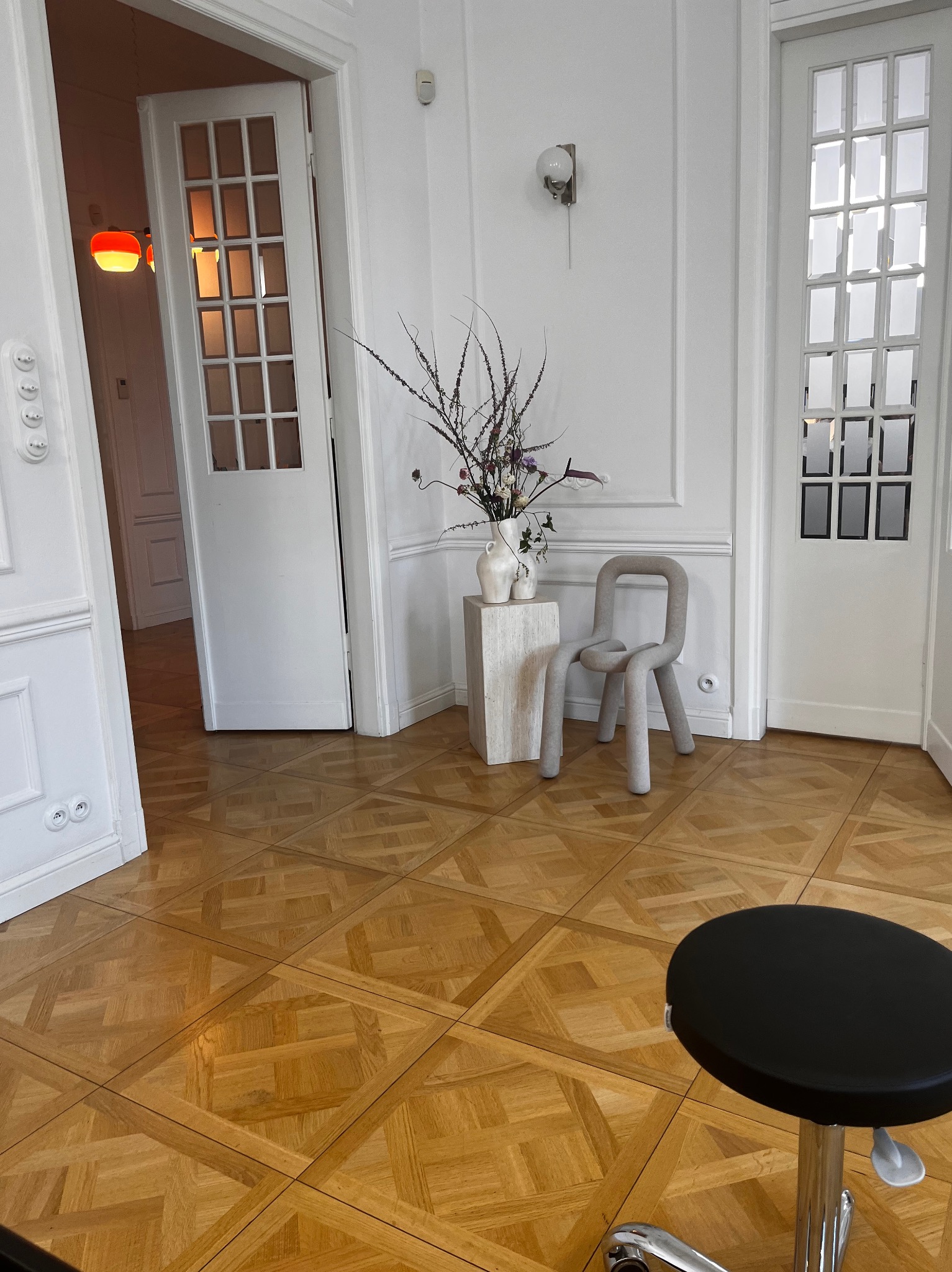 Jasne wnętrze z parkietem w jodełkę, białymi drzwiami z przeszkleniami i sztukaterią, nowoczesnym krzesłem z szarej tkaniny, wazonami z kwiatami na postumencie i czarnym stołkiem na kółkach