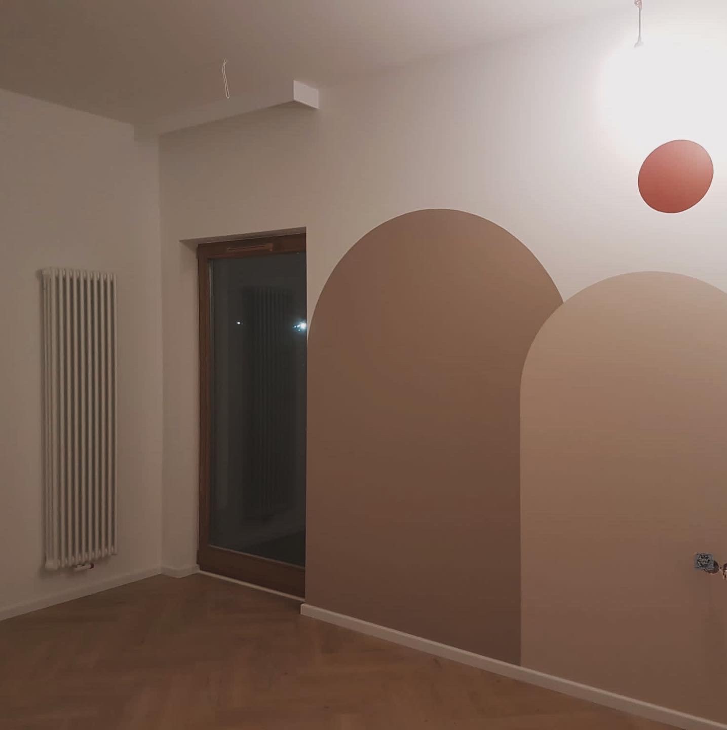 Pomalowane ściany w pokoju z dekoracyjnymi łukami w odcieniach brązu, drewniana podłoga, grzejnik i okno z drewnianą ramą.