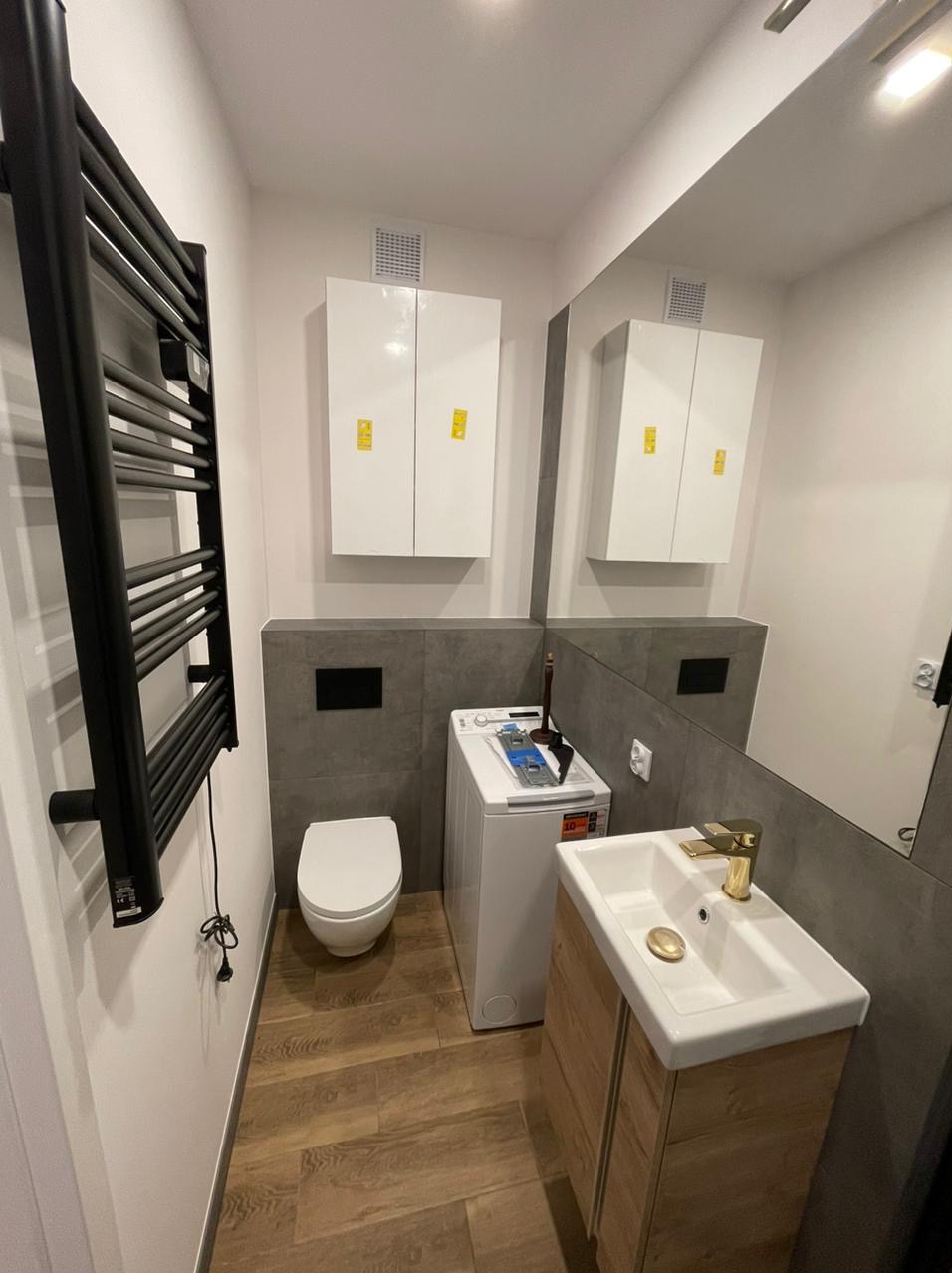 Nowoczesna łazienka z drewnopodobną podłogą, szarą glazurą, białym WC i szafkami, pralką oraz umywalką z złotą armaturą.