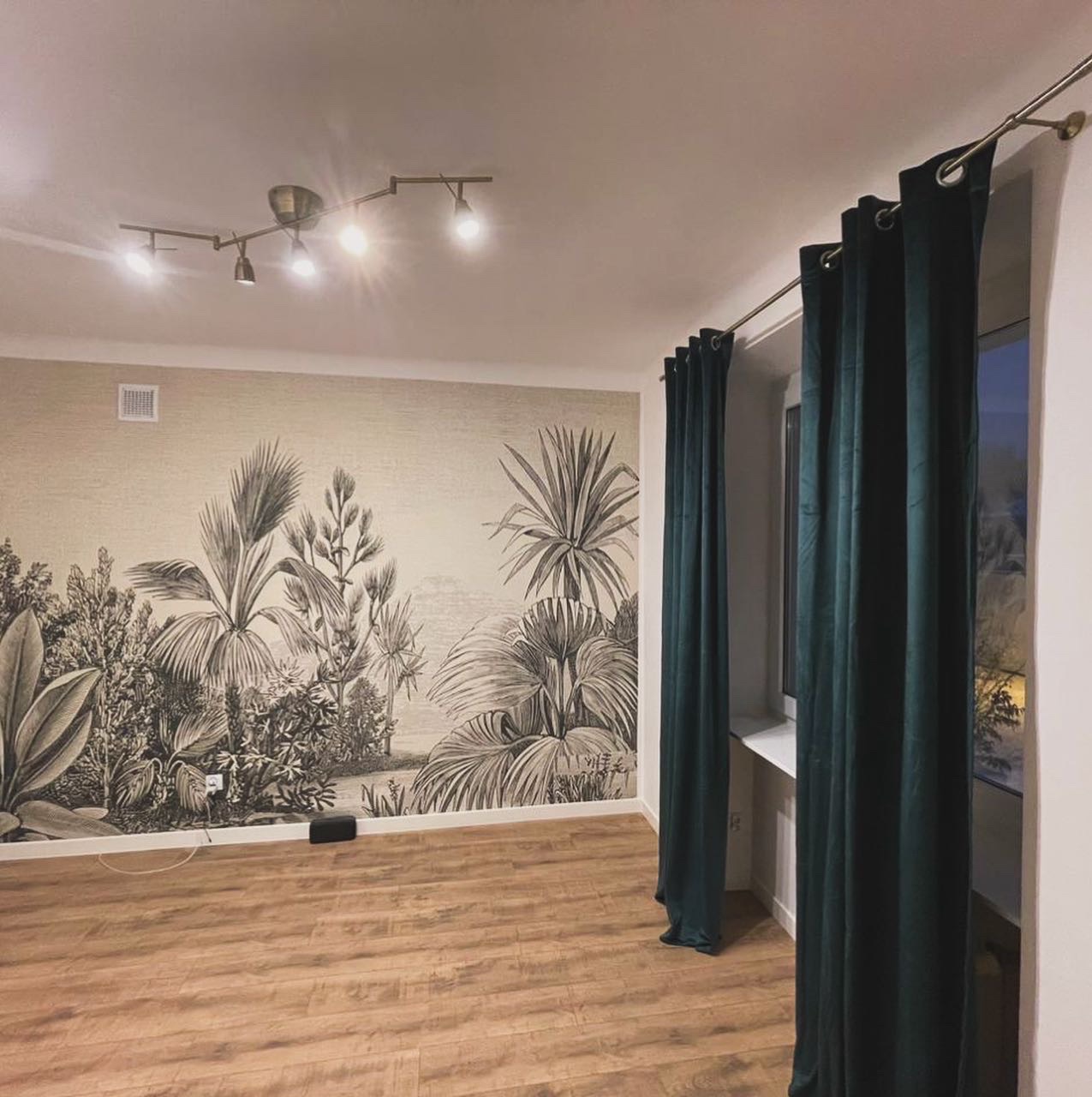 Pokój po remoncie z fototapetą w stylu botanicznym, drewnianą podłogą i ciemnozielonymi, welurowymi zasłonami przy oknie.