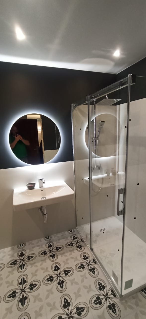 Nowoczesna łazienka z kabiną prysznicową typu walk-in, okrągłym lustrem z podświetleniem LED, wiszącą umywalką z miseczką na mydło i wzorzystą podłogą w odcieniach szarości.