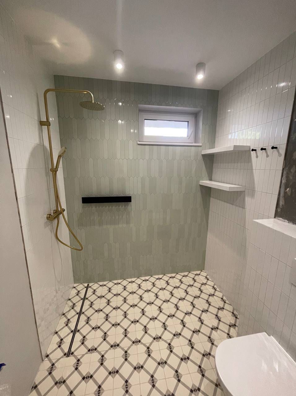 Wykończona łazienka w Nowej Woli z kabiną prysznicową z złotą armaturą, geometryczną podłogą w romby i pionowymi płytkami w odcieniach szarości na ścianach, uzupełniona o białe półki i okno.