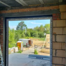 Montaż okna PCV w surowym stanie, widok z wnętrza budynku na podwórze z białym dostawczakiem i składowanym drewnem, widoczne elementy rusztowania i drabina.