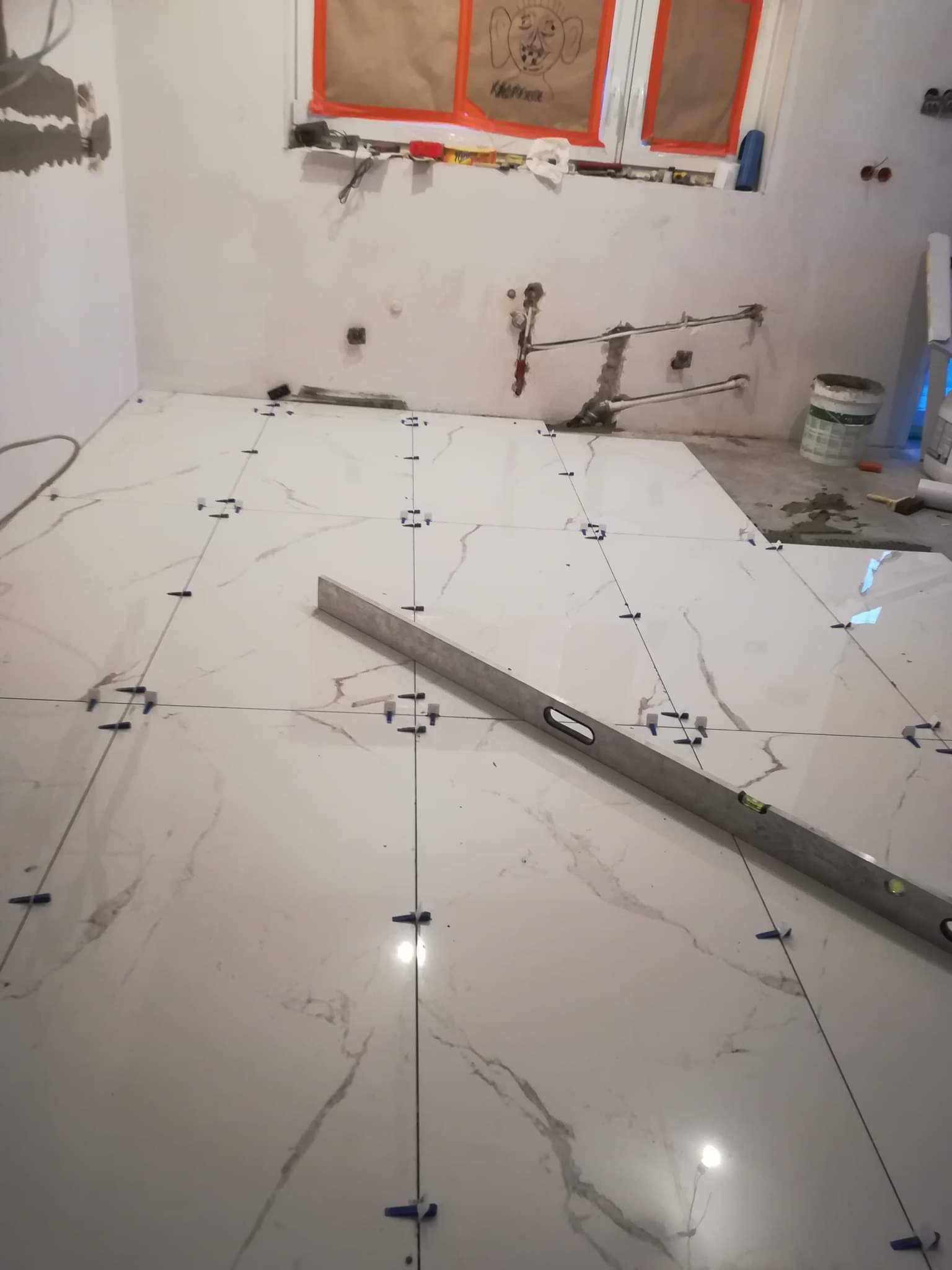 Układanie białych, marmurkowych płytek podłogowych z użyciem krzyżyków dystansowych i poziomicy w trakcie prac wykończeniowych w łazience.