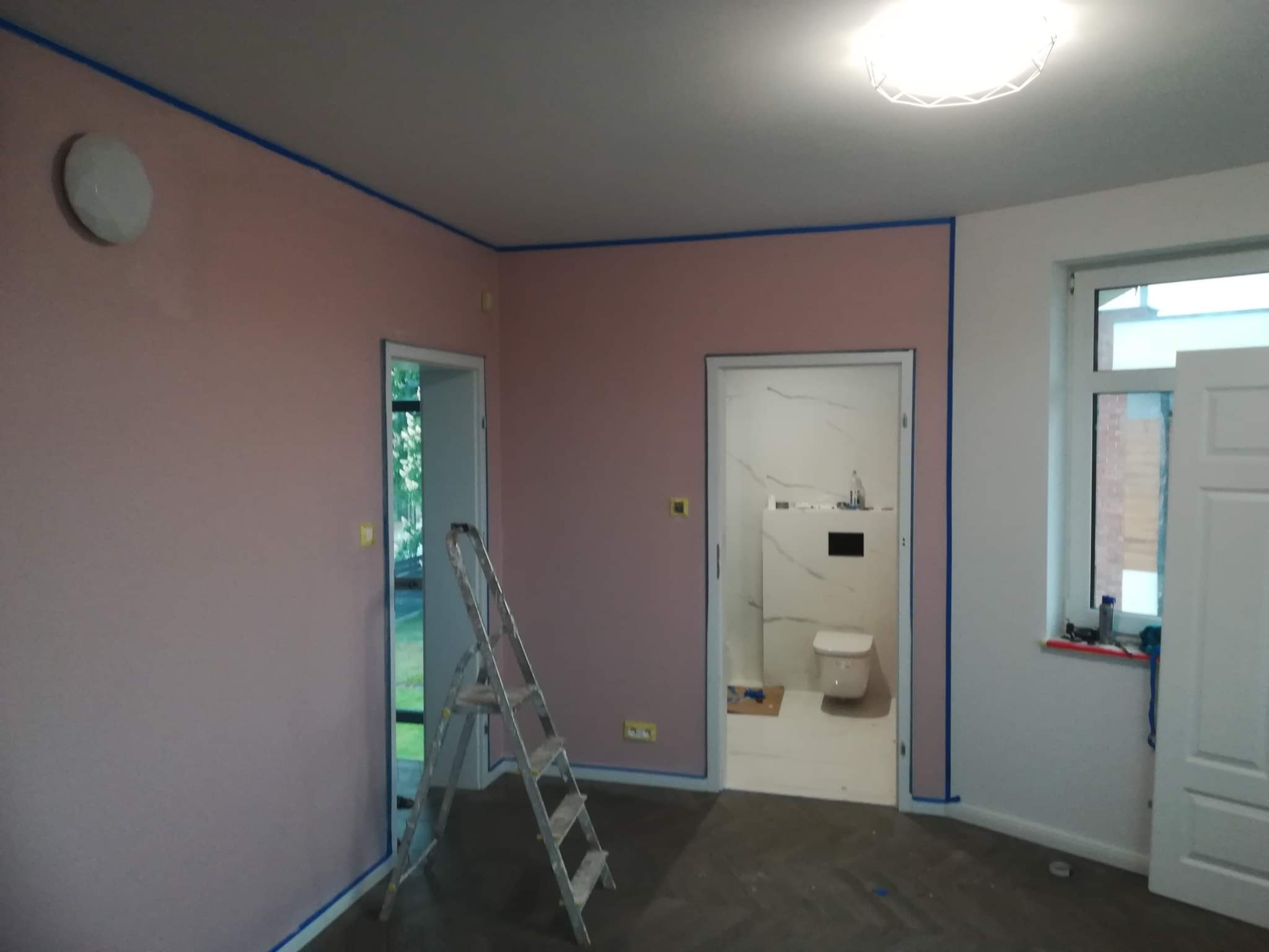 Pokój z różowymi ścianami w trakcie przygotowania do malowania, zabezpieczone niebieską taśmą, widoczna drabina aluminiowa i fragment łazienki z białymi płytkami.