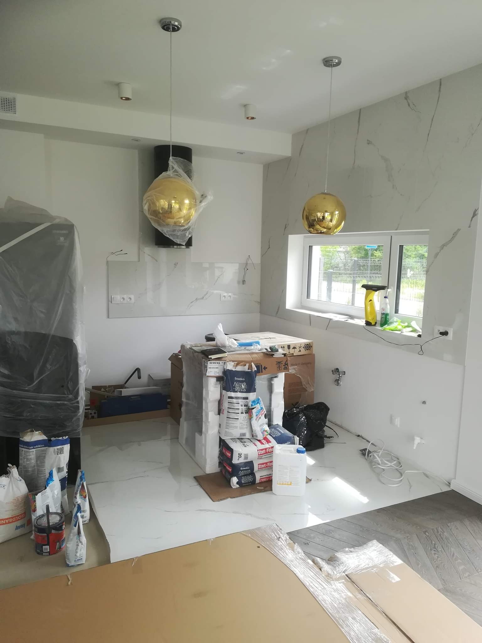 Wnętrze kuchni w trakcie prac wykończeniowych: marmurowe ściany i podłogi, złote lampy wiszące w foliach ochronnych, kartony i worki z materiałami budowlanymi, zakryta folią lodówka, widoczne...