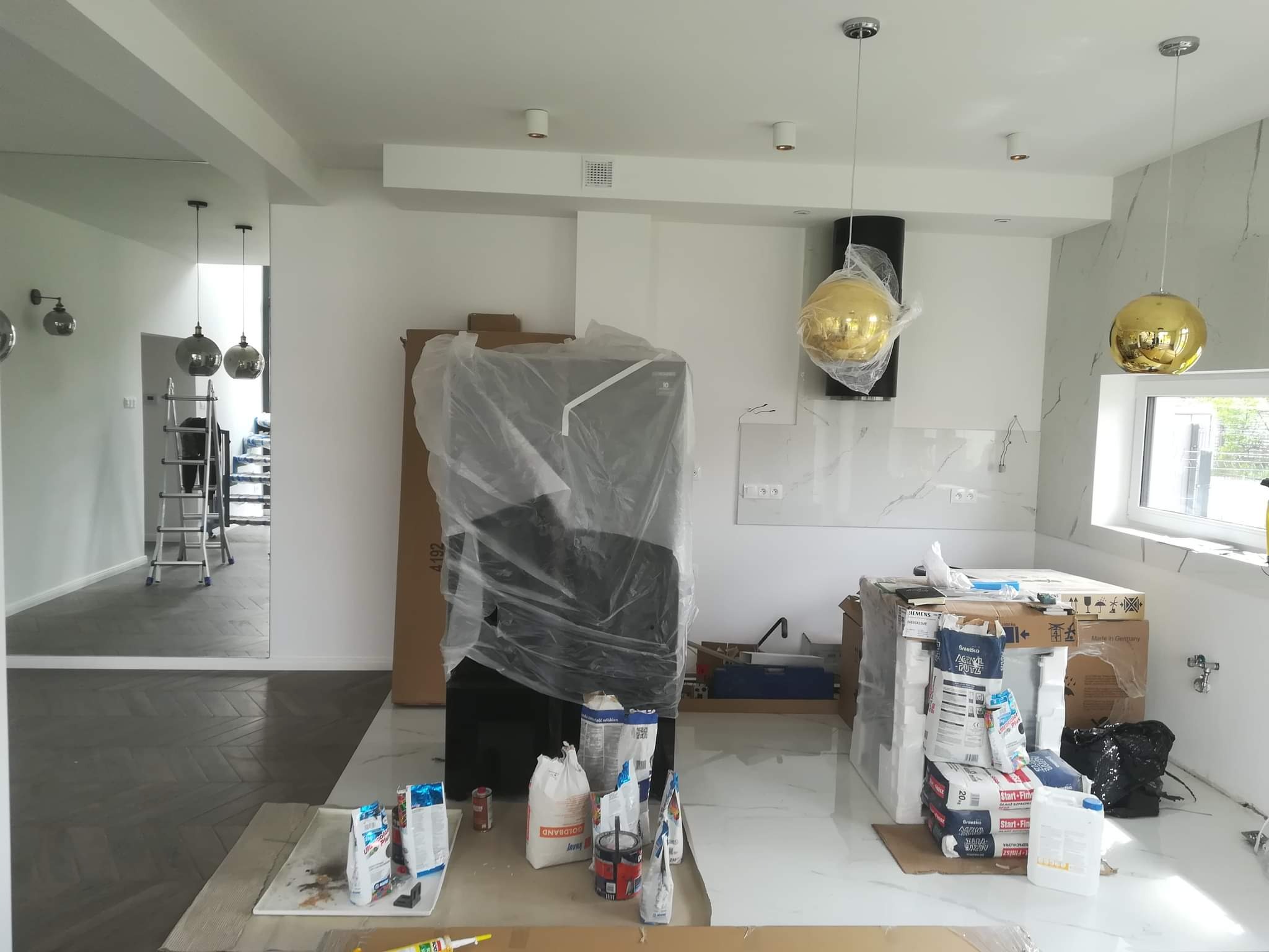 Wnętrze kuchni w trakcie prac wykończeniowych: podłoga wyłożona płytkami imitującymi marmur, widoczne worki z gładzią i narzędzia, sprzęt AGD zabezpieczony folią, złote lampy wiszące.