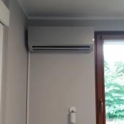 Szary klimatyzator Rotenso zamontowany na ścianie w pokoju, widok z okna na zewnątrz.