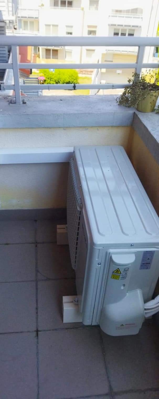 Zewnętrzna jednostka klimatyzatora zamontowana na balkonie, widoczne białe wsporniki i kable, w tle zabudowa wielorodzinna.