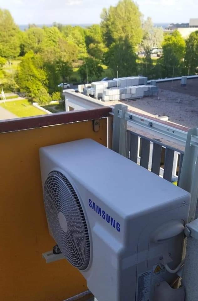 Zewnętrzna jednostka klimatyzatora Samsung zamontowana na balkonie z widokiem na zielone drzewa i budynki w tle.