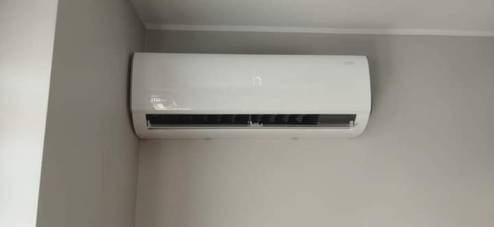 Biała jednostka klimatyzacyjna zamontowana na szarej ścianie w pomieszczeniu, widoczny wyświetlacz temperatury.