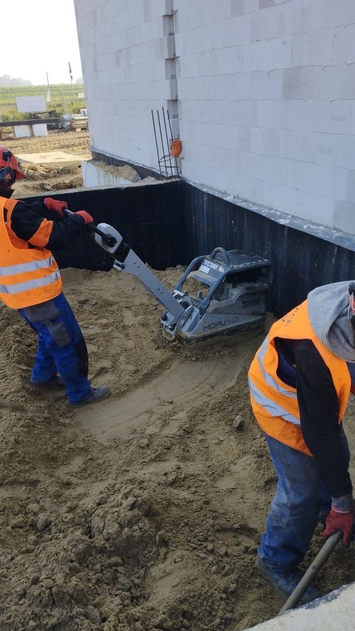 Prace ziemne: Zagęszczanie piasku zagęszczarką wibracyjną przy fundamencie powstającego budynku z białego bloczka.